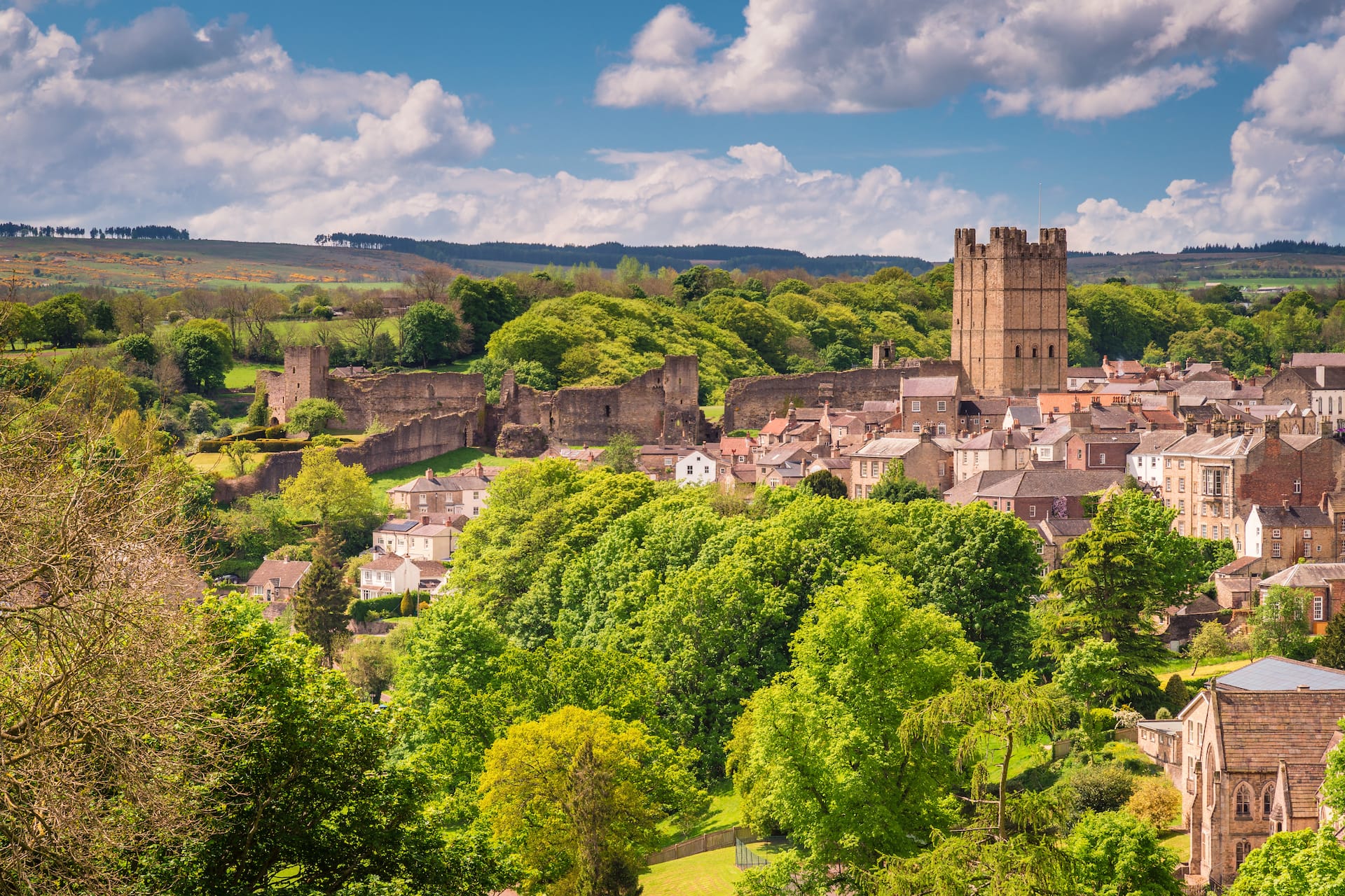 Historic English town with castle ruins, dense green trees, and rolling hills under a blue sky.