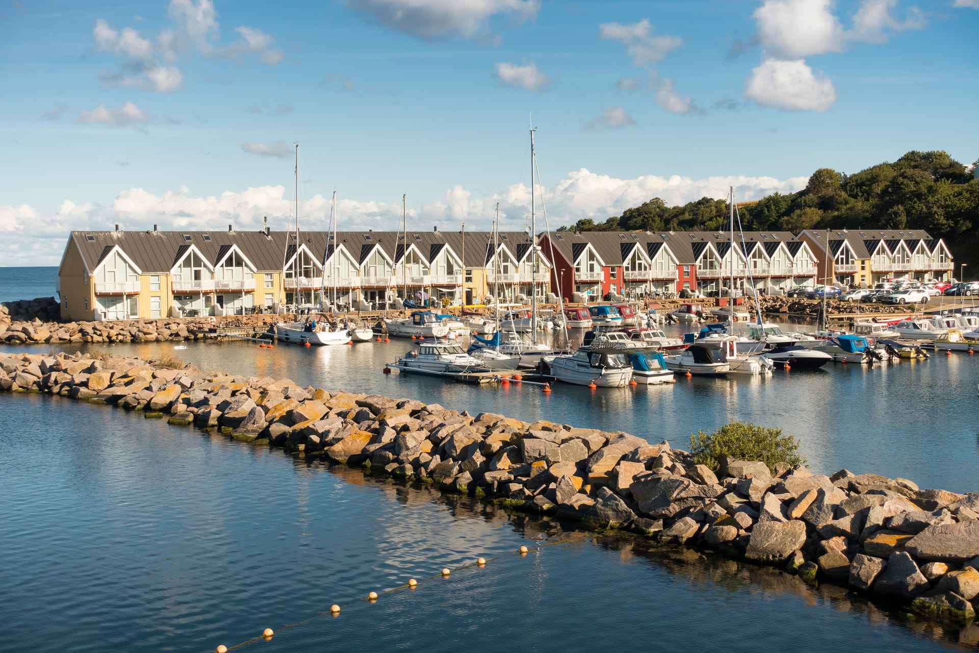 Boats docked in a harbor with yellow waterfront houses and a rock jetty under blue sky.