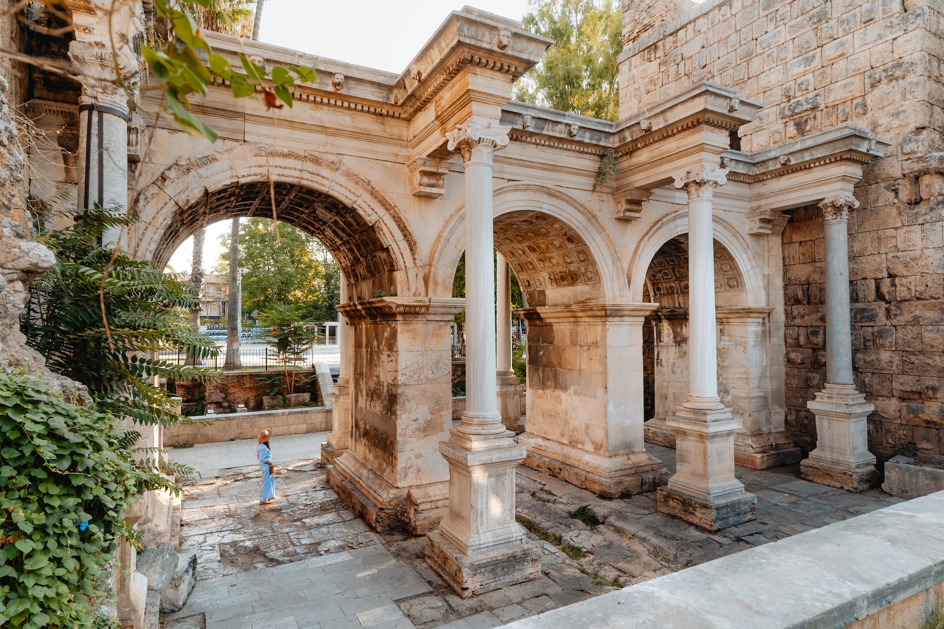 Ancient stone triumphal arch with columns, woman in sun hat standing below foliage.