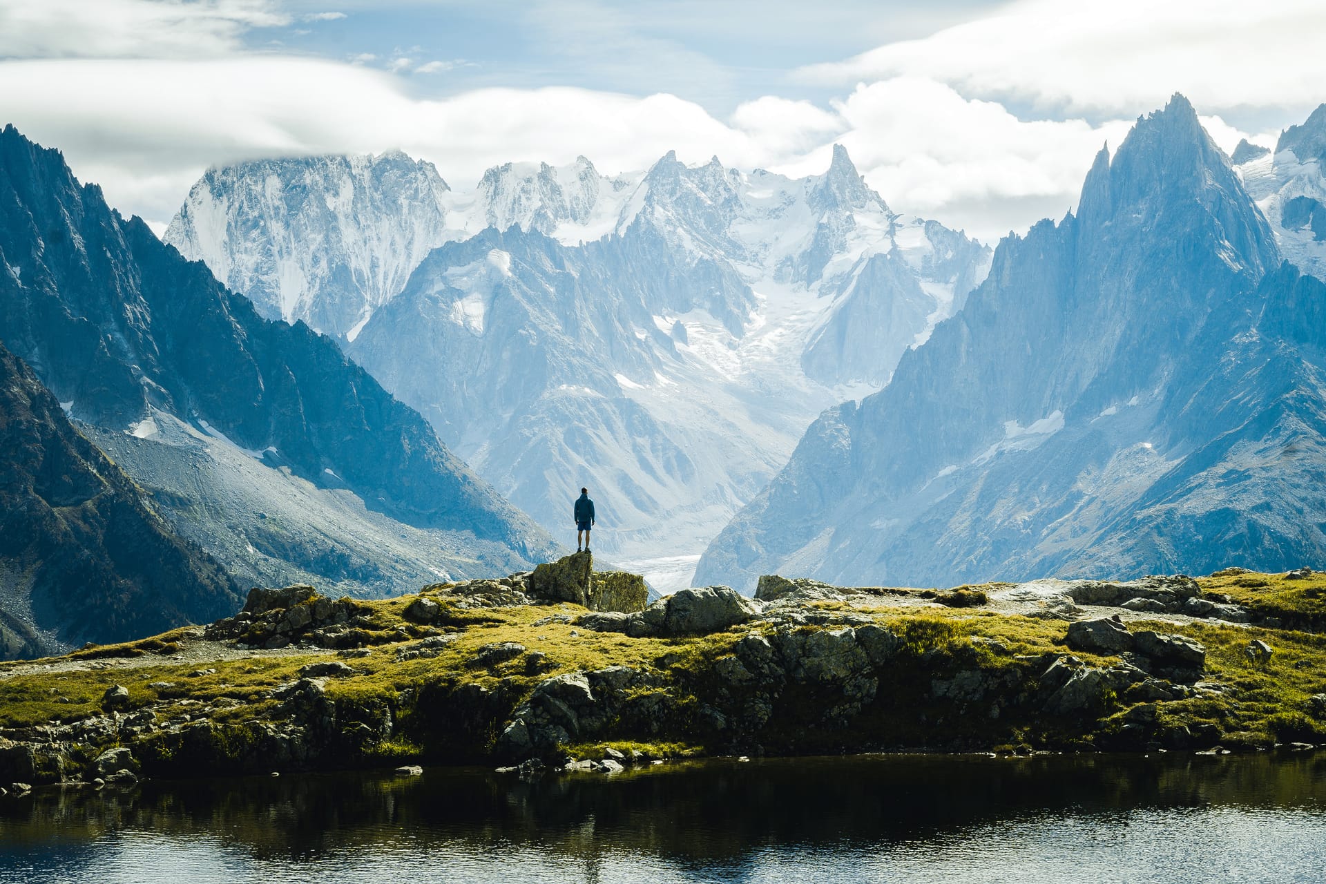 Hiker standing on rocky outcrop overlooking massive snow-capped alpine mountains and lake.