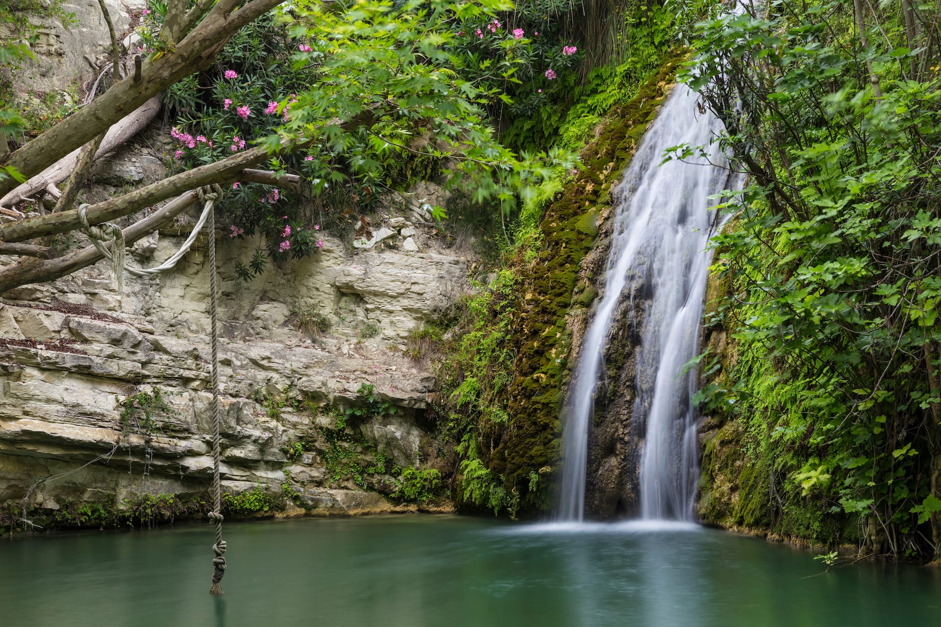 Waterfall cascading into a green pool with a rope swing by mossy cliffs in Cyprus.