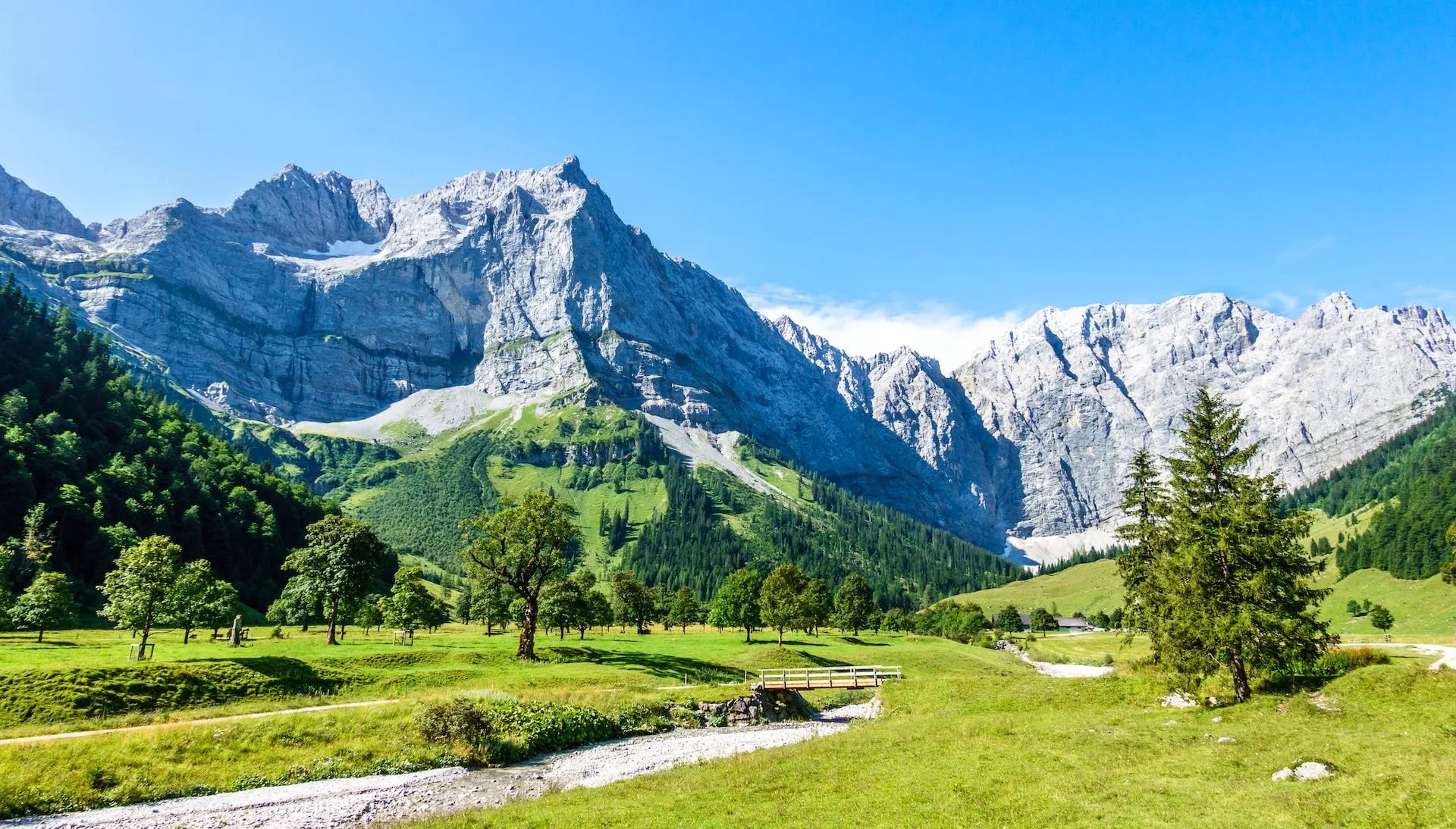 Steep rock faces and green meadows with a small bridge over a stream in the mountains