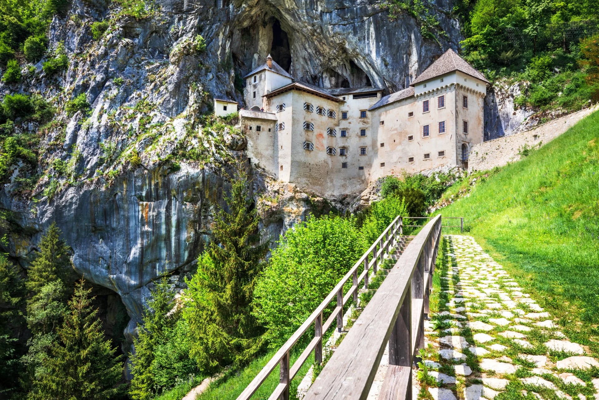 Castle built into a cliff face above a stone path leading uphill through lush green summer foliage.