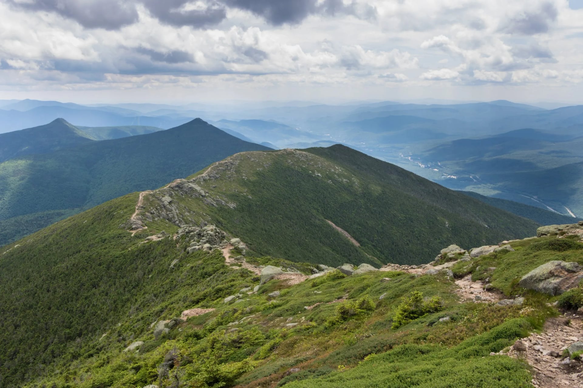 A worn trail running along an open grassy crest with peaks beyond