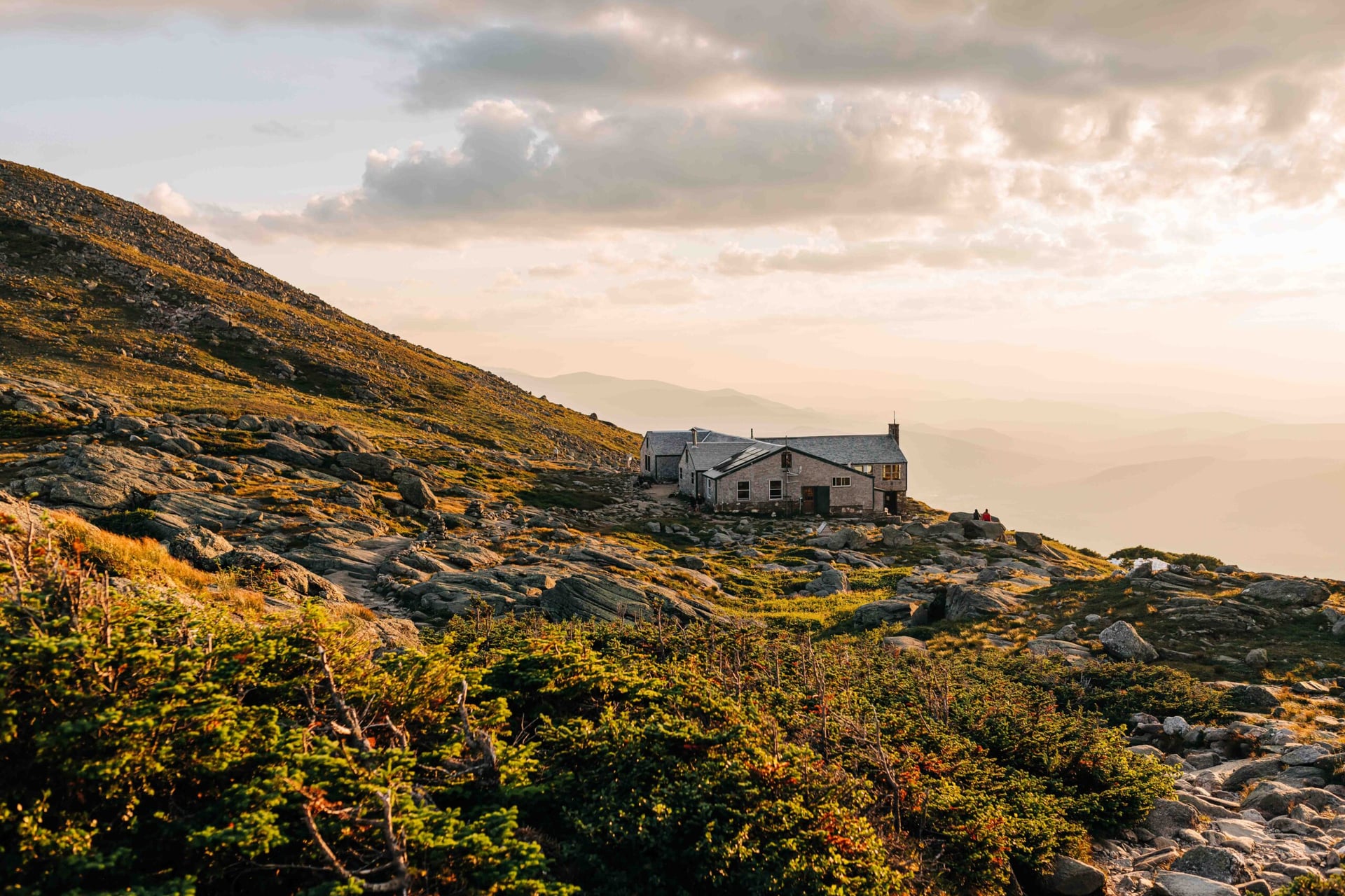 A stone hut on a rocky slope of low scrub with a sea of cloud below