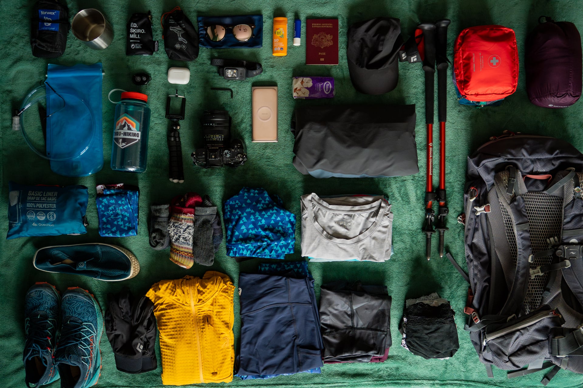 Hiking and trekking gear laid out on a green surface, including a backpack, trekking poles, camera, and clothing.