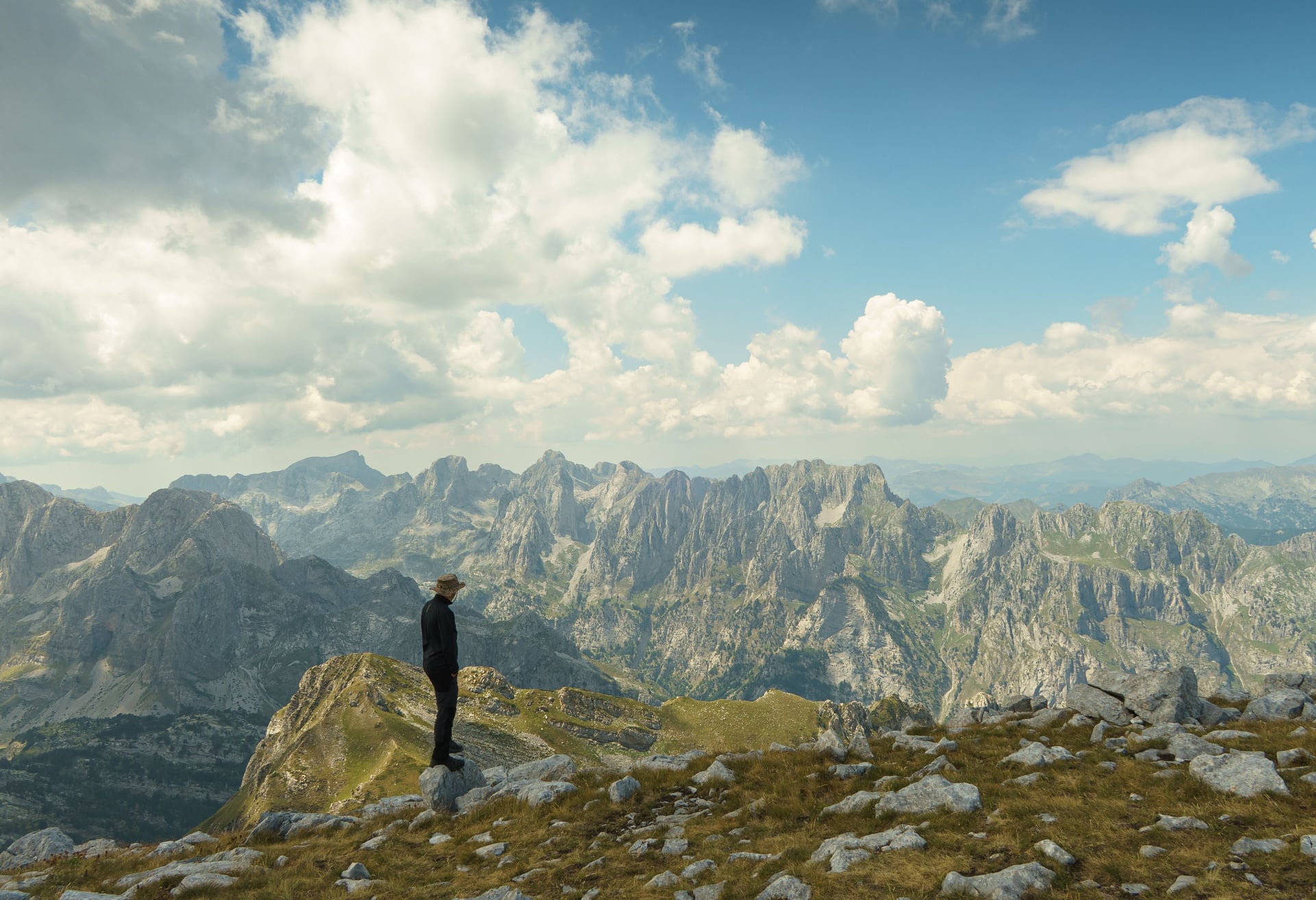 Person standing on a rocky mountain peak overlooking a vast, rugged mountain range under a cloudy sky.