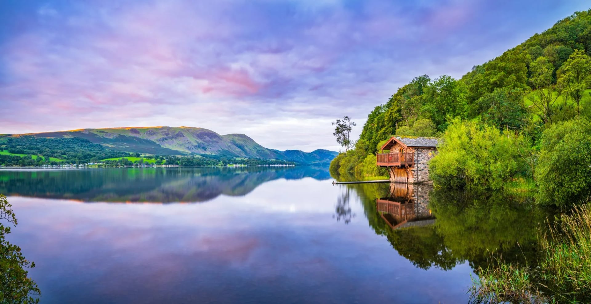 A boathouse on the still water of Ullswater under a pink dawn sky