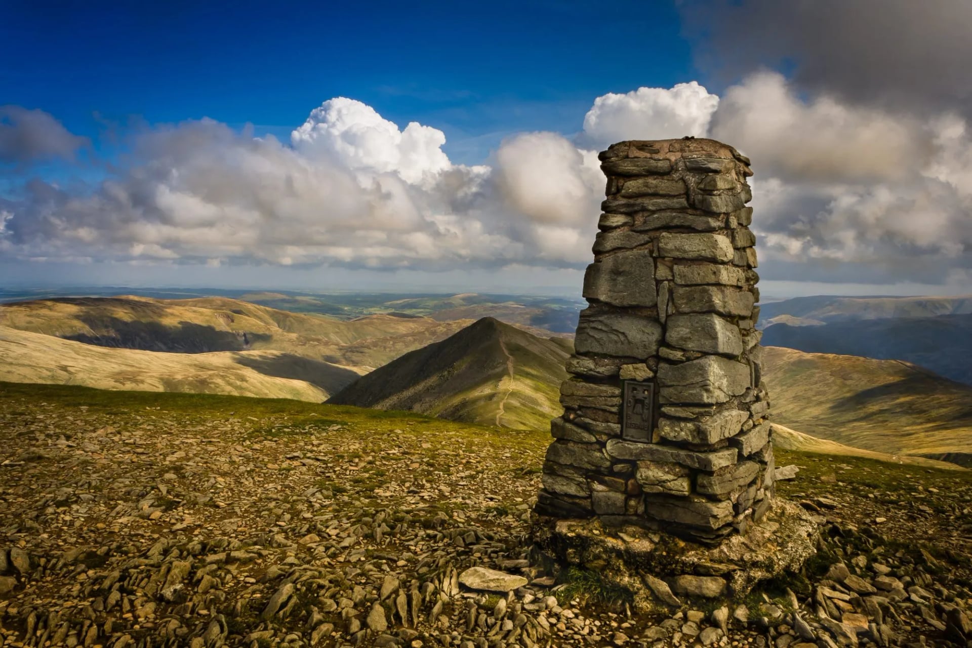 A stone trig pillar on a stony summit with fells and a lake beyond