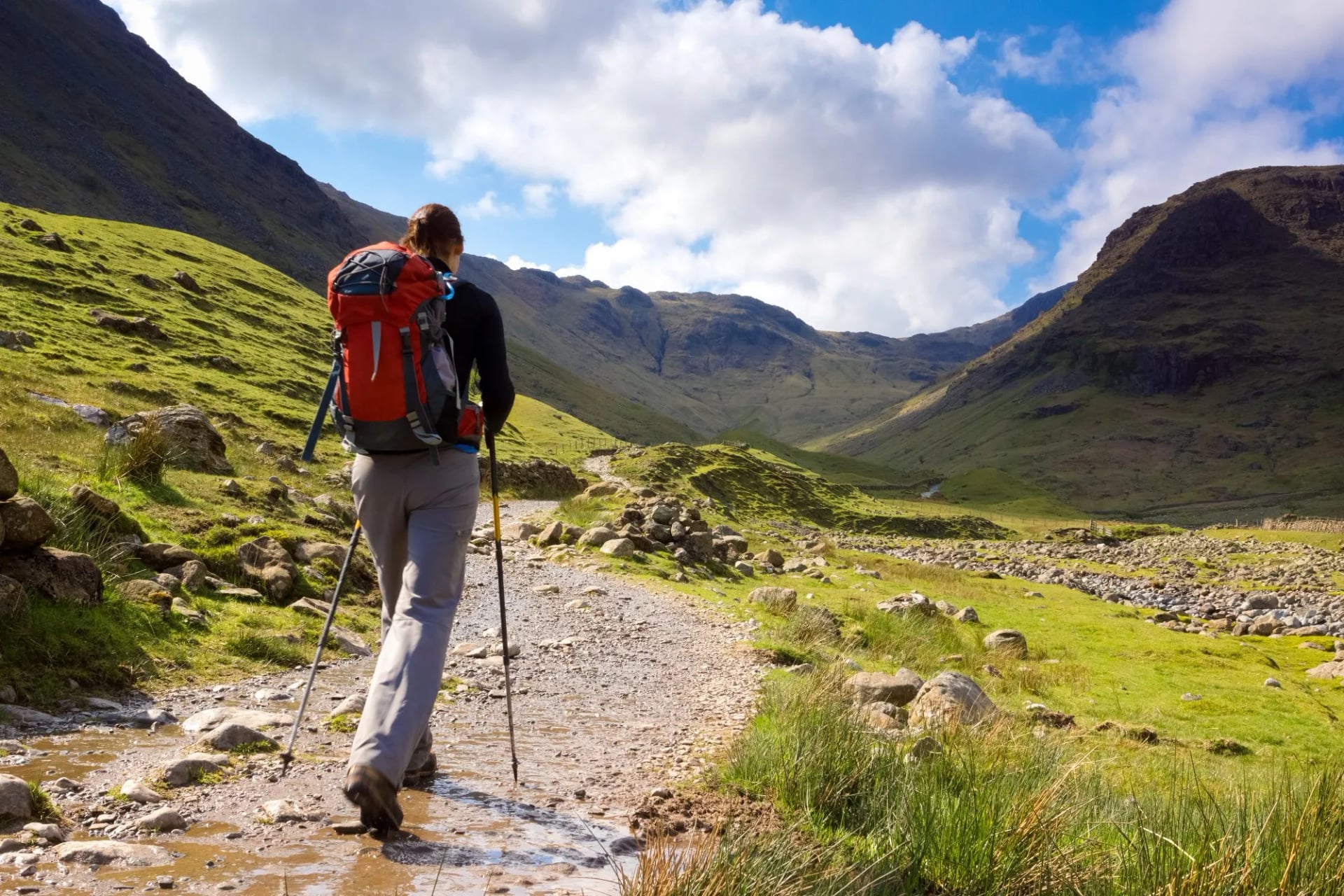 A walker with poles and a red pack on a stony path in a green Lakeland valley