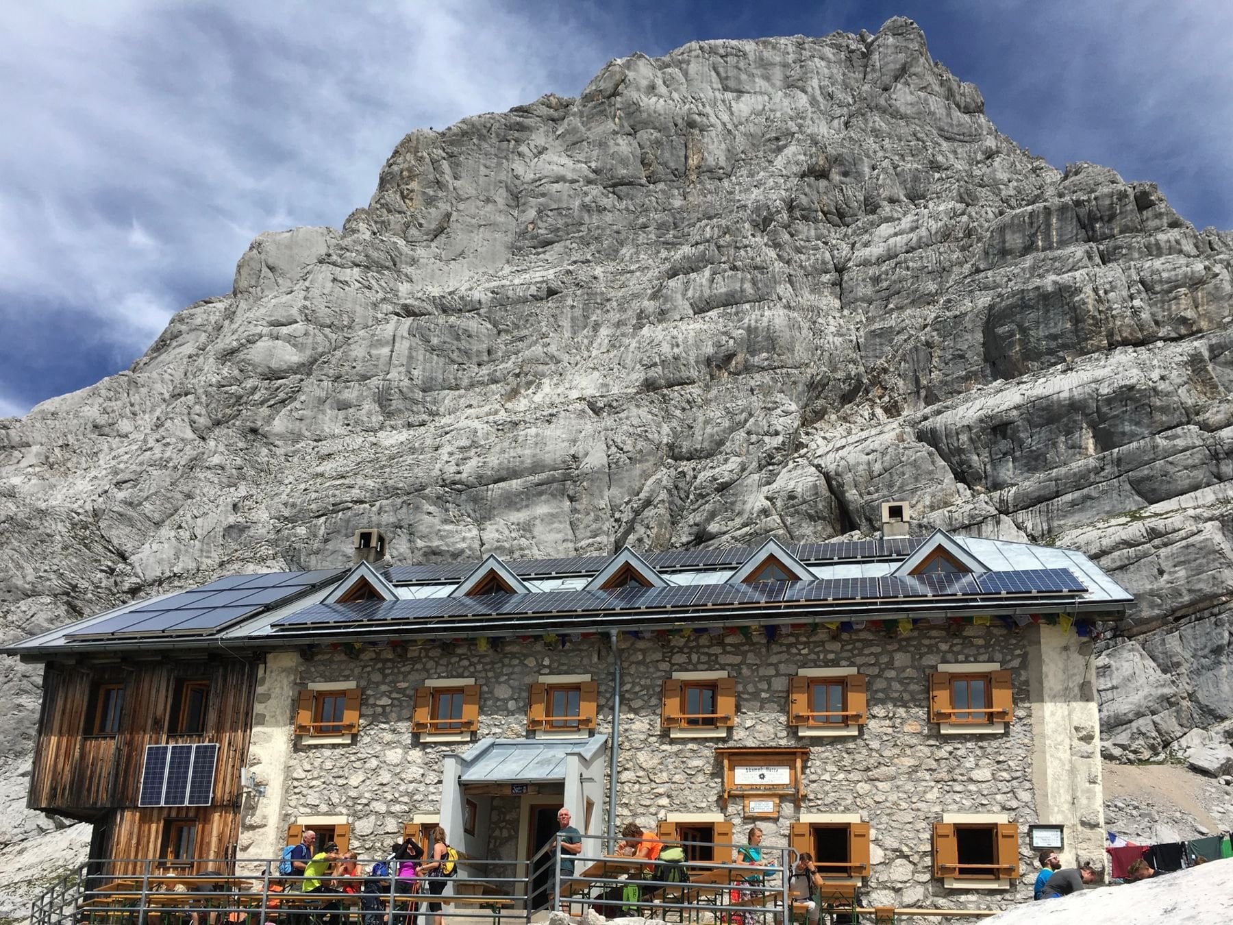 A stone mountain hut with walkers outside below rock faces