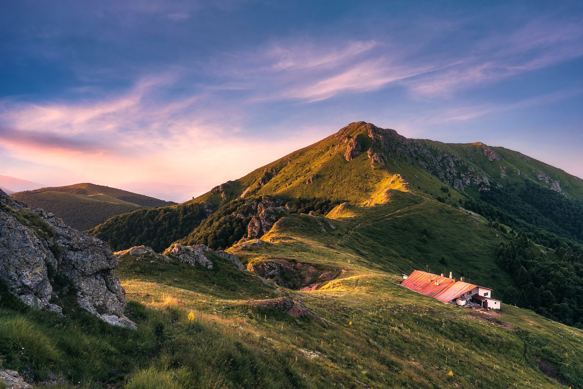 Mountain landscape with grassy hills and a small building under a sunset sky