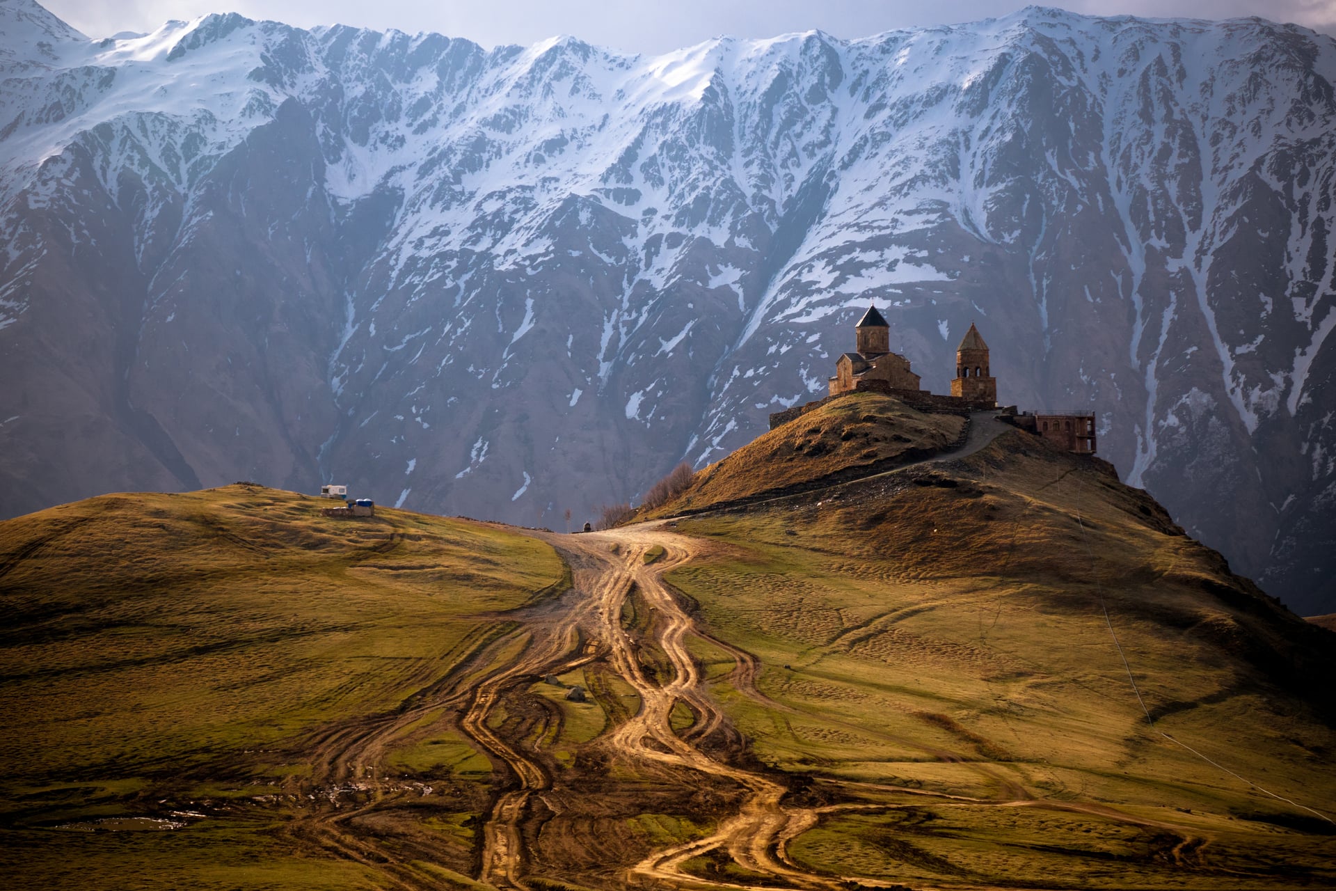 Gergeti Trinity Church on hill with dirt tracks and snow-covered mountain backdrop in Georgia