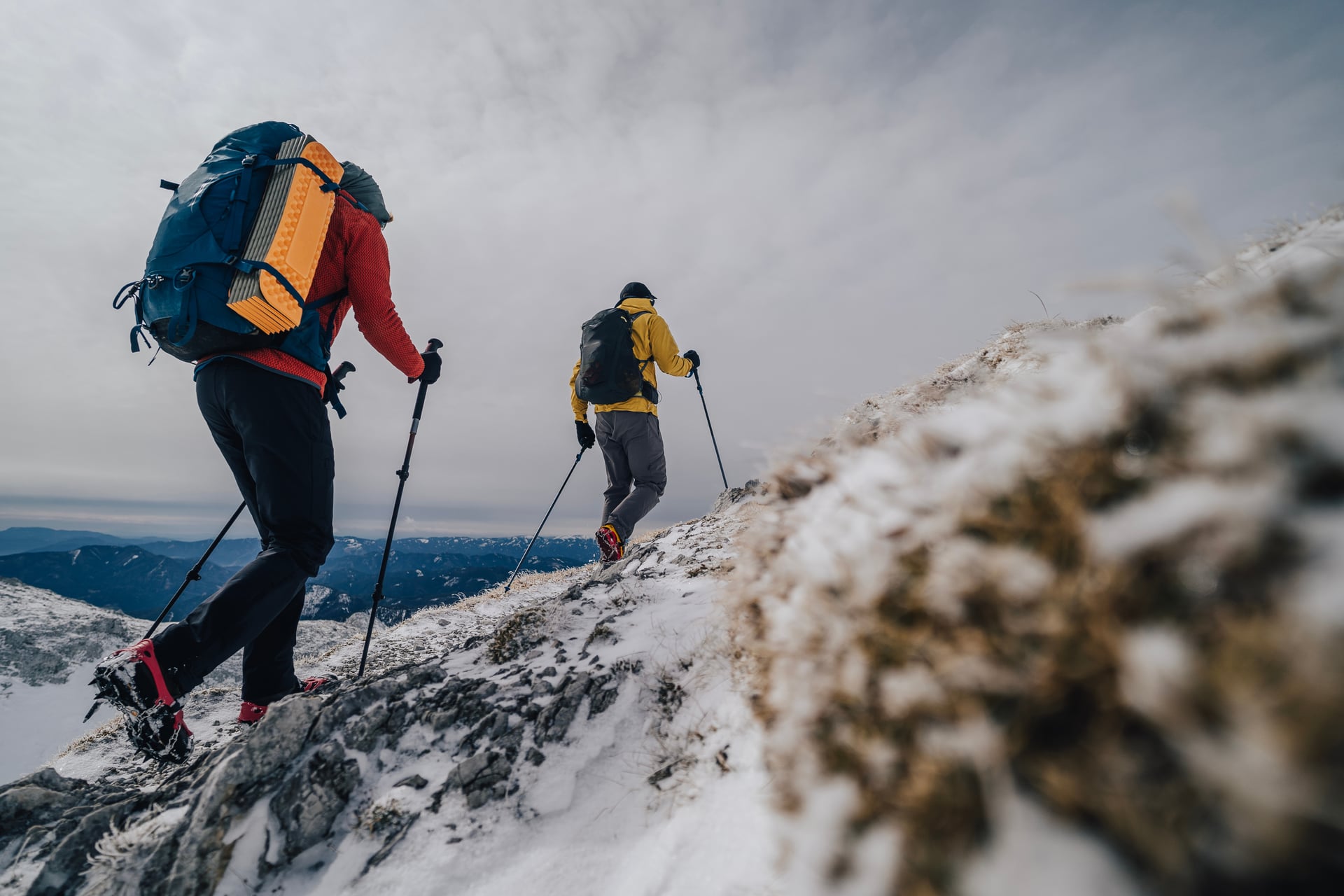 Två vandrare med ryggsäckar och vandringsstavar som går uppför en snöklädd bergskam