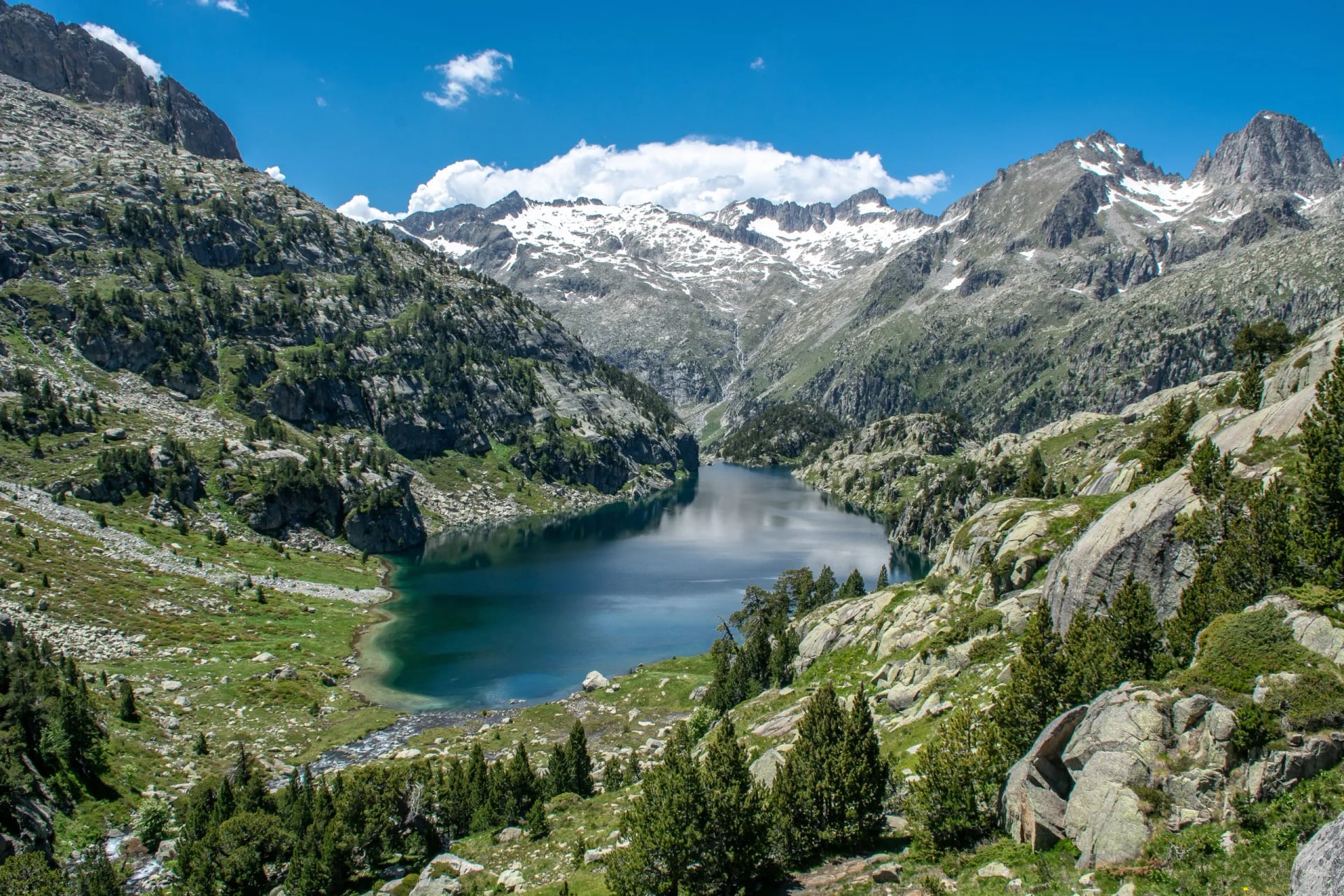 Deep blue lake ringed by pine forest below snow streaked granite peaks in Aigüestortes
