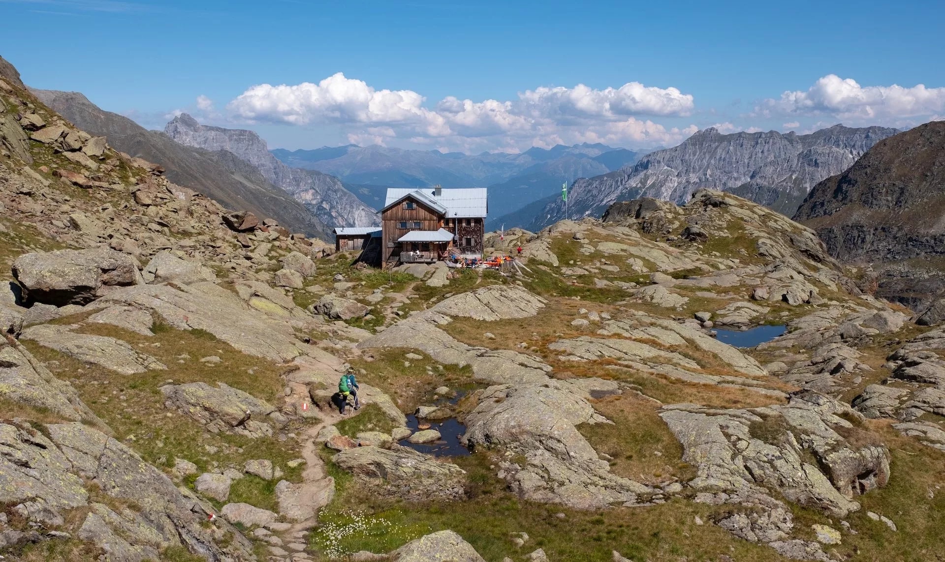 Alpine hut on rocky terrain with hiker approaching, surrounded by mountains under blue sky.