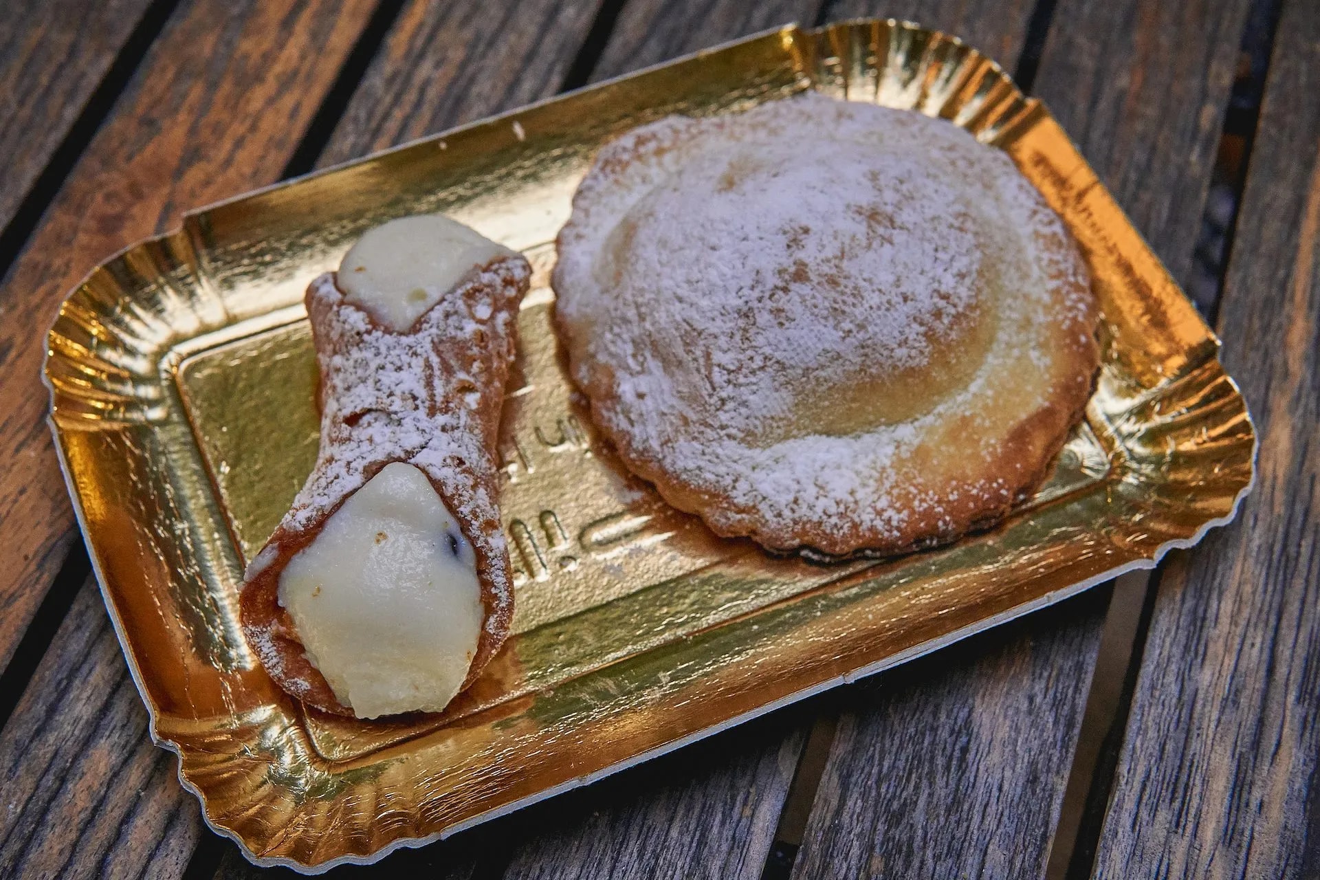 Cannoli and pastry dusted with powdered sugar on a gold tray on wood