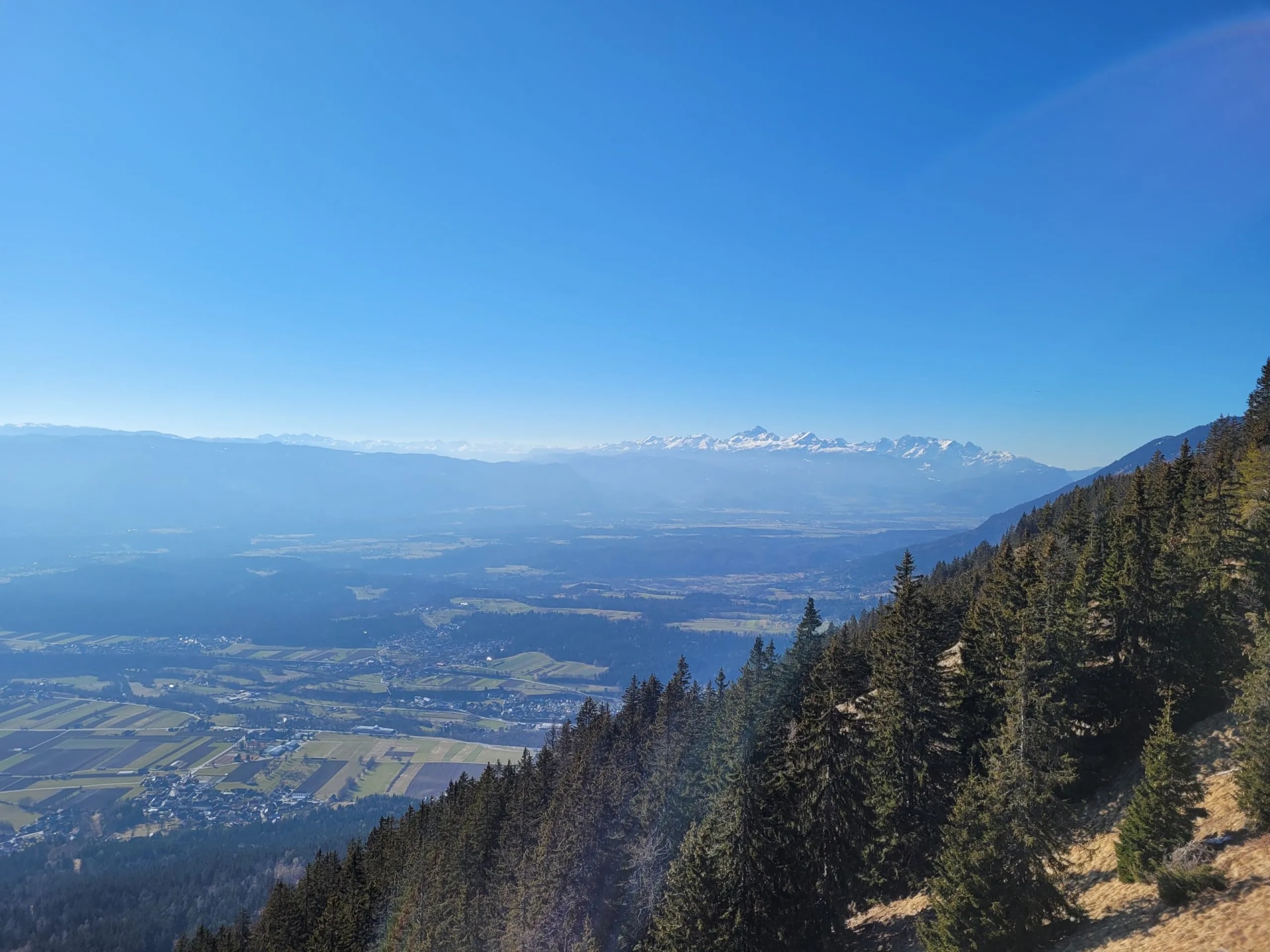 Mountain view overlooking valley with village and distant snow-capped peaks under clear blue sky.