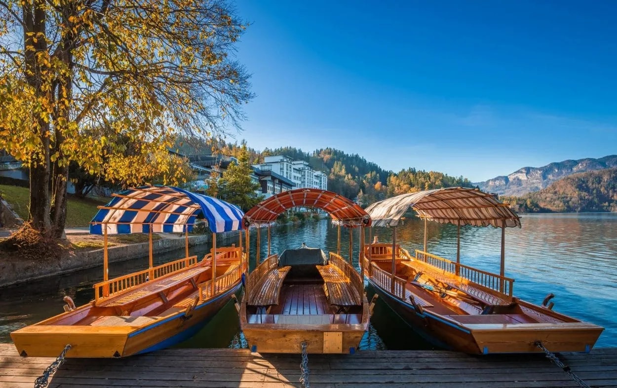 Three traditional wooden Pletna boats docked on a pier at Lake Bled with autumn foliage and mountains.