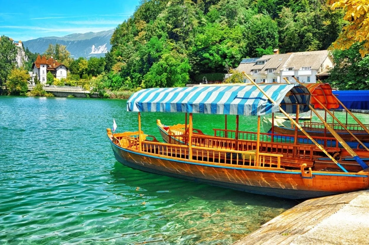 Pletna boat docked on emerald water of Lake Bled with forested mountains and houses.