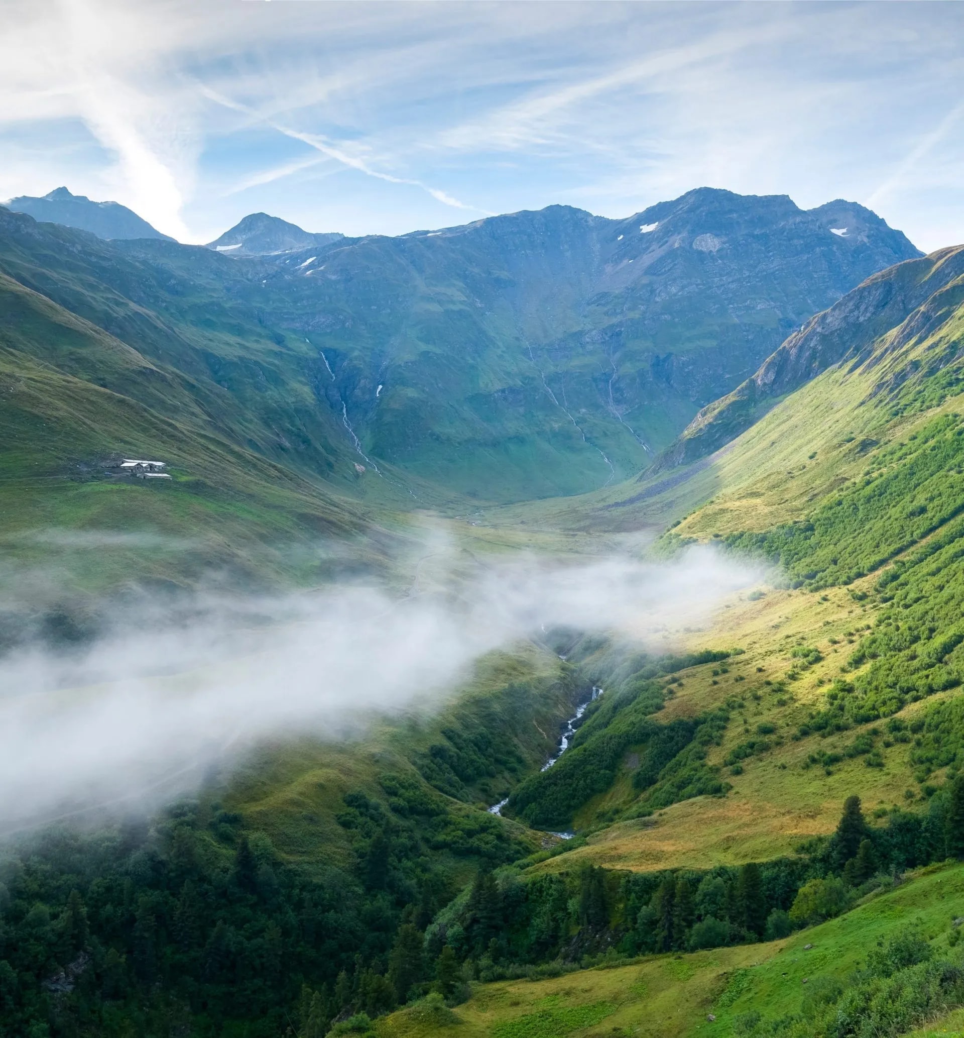 Green mountain valley with low fog, small buildings, and waterfalls under a blue sky.