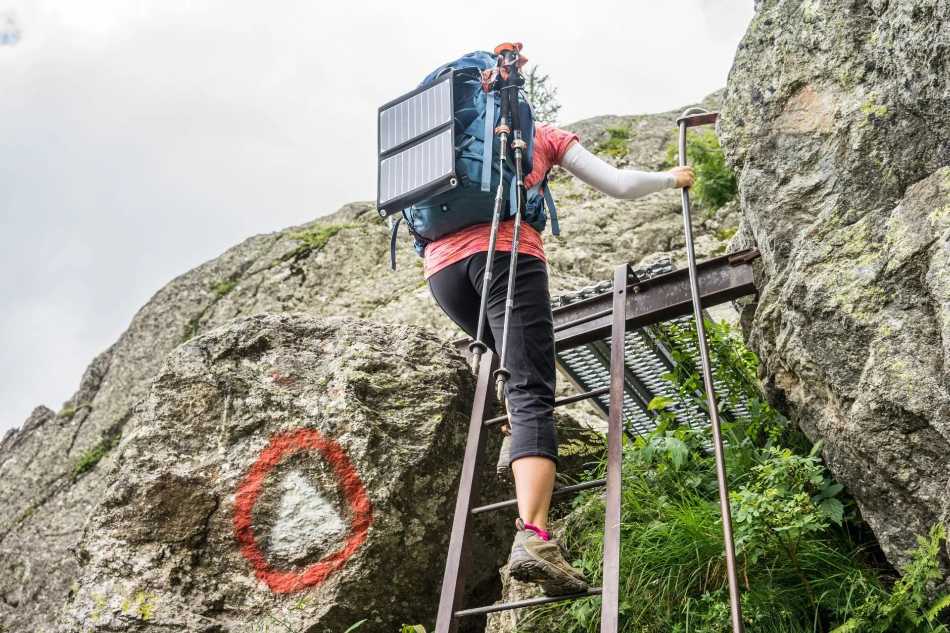 Hiker with solar panel backpack ascending metal ladder on rocky mountain trail with trail marker.