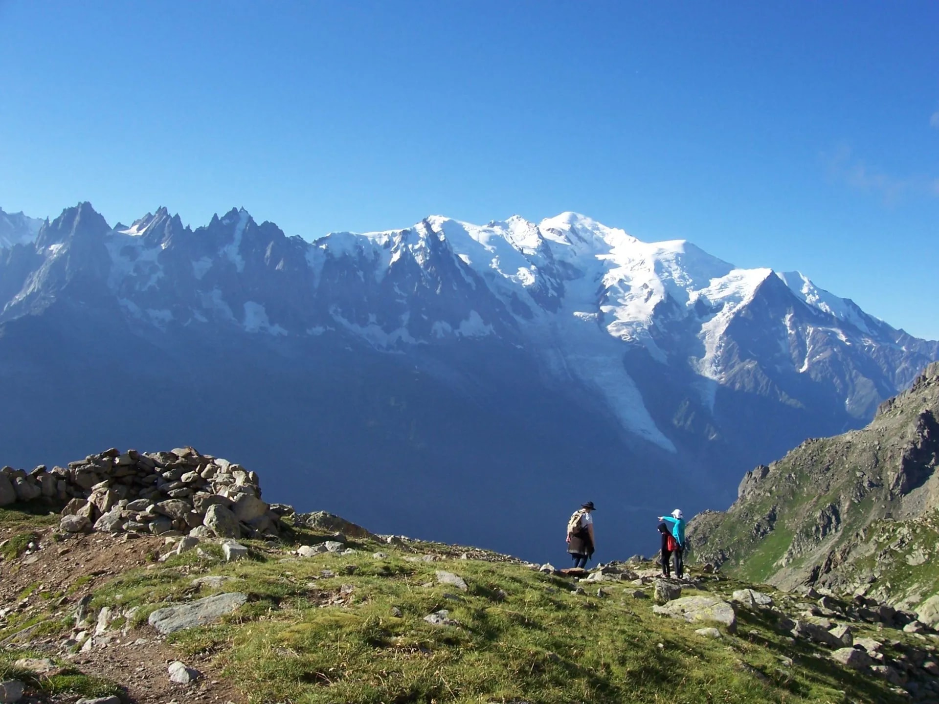 Hikers on grassy ridge overlooking snow-capped mountains, likely Le Brevent range.