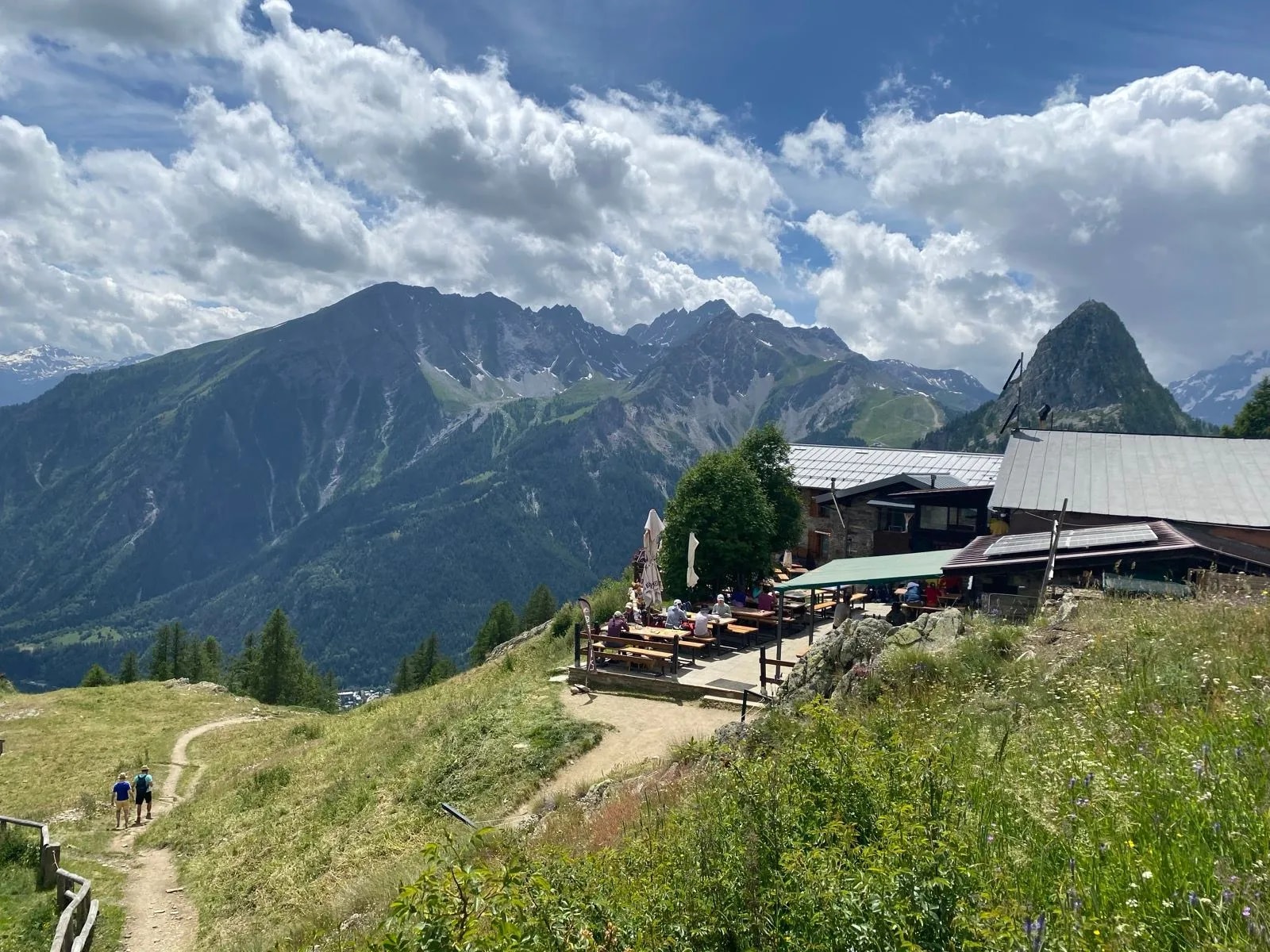 Hikers on trail near mountain refuge with outdoor dining and dramatic peaks under cloudy sky