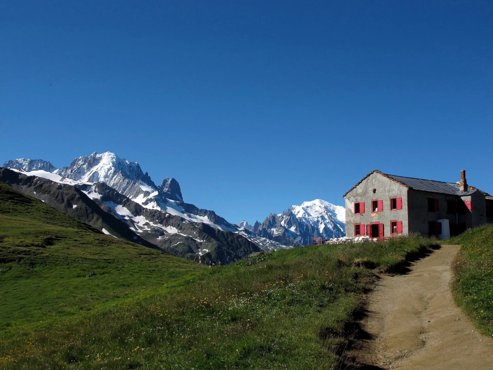 Stone refuge hut with red shutters on a grassy slope near snow-capped mountains at Col de Balme.