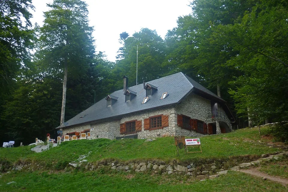 Stone mountain refuge Rifugio di Conangles surrounded by dense green forest with outdoor seating area.