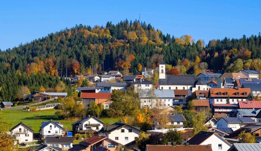 Alpine village houses nestled below a forest with autumn foliage in Bavaria under a clear blue sky.
