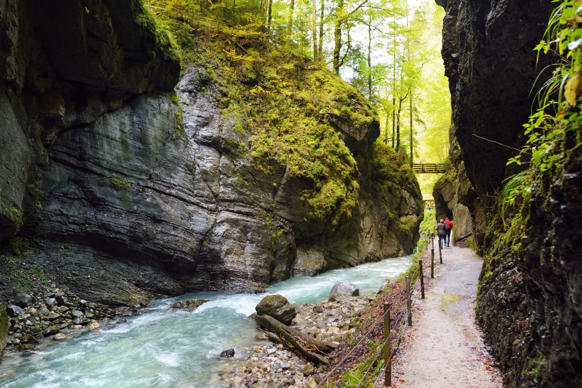 Hikers on a path beside a turquoise river through the mossy, narrow Partnach Gorge.