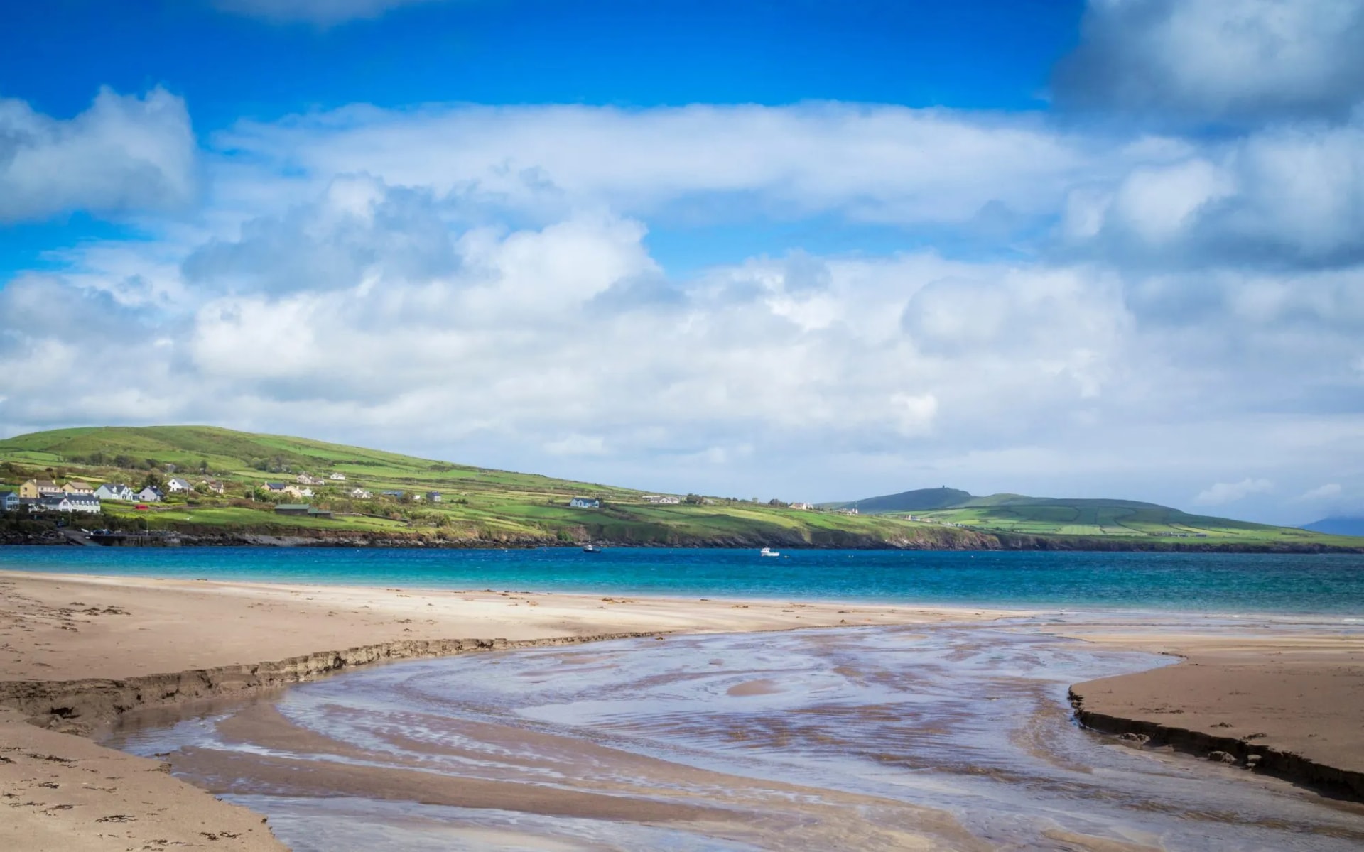 Sandy beach with stream flowing to turquoise water, green hills, and Ventry village.