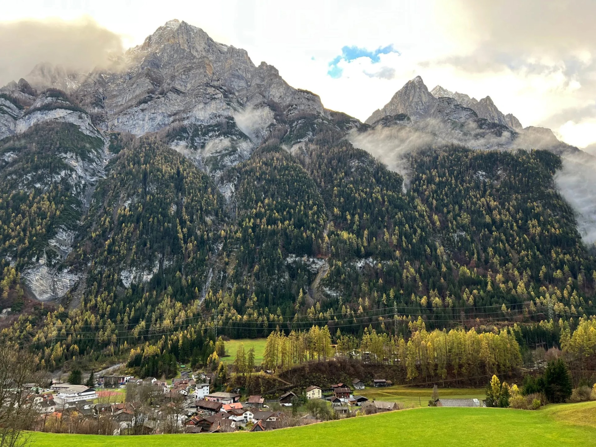 Alpine village nestled below steep, forested mountains with autumn foliage and low clouds near Haldensteiner Calanda.