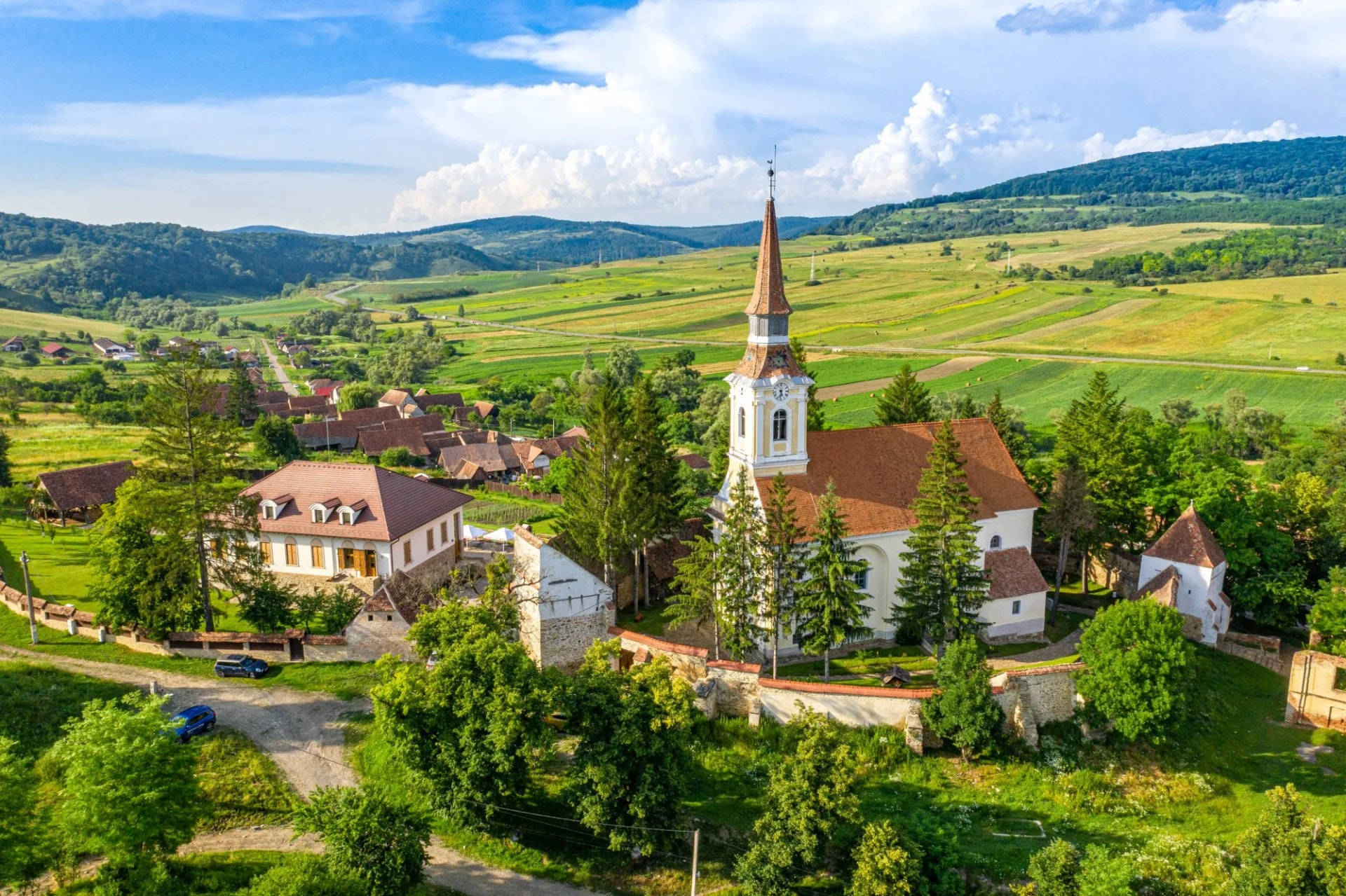 Church with tall spire in small village surrounded by green rolling hills under blue sky.
