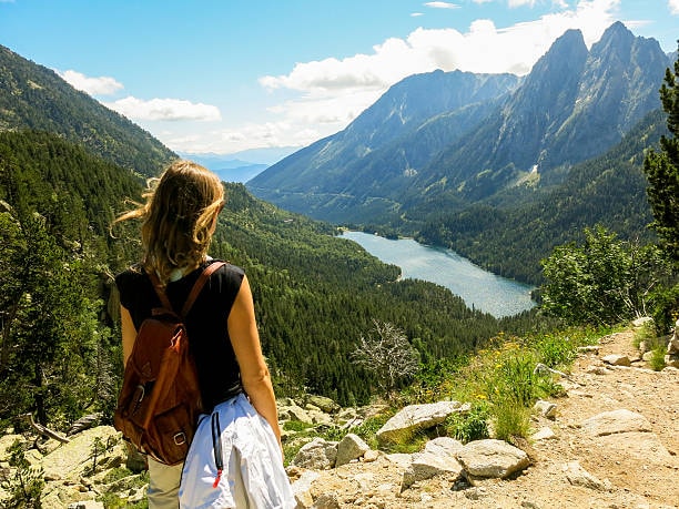 Hiker with a backpack looking out over an alpine lake surrounded by mountains and forest.