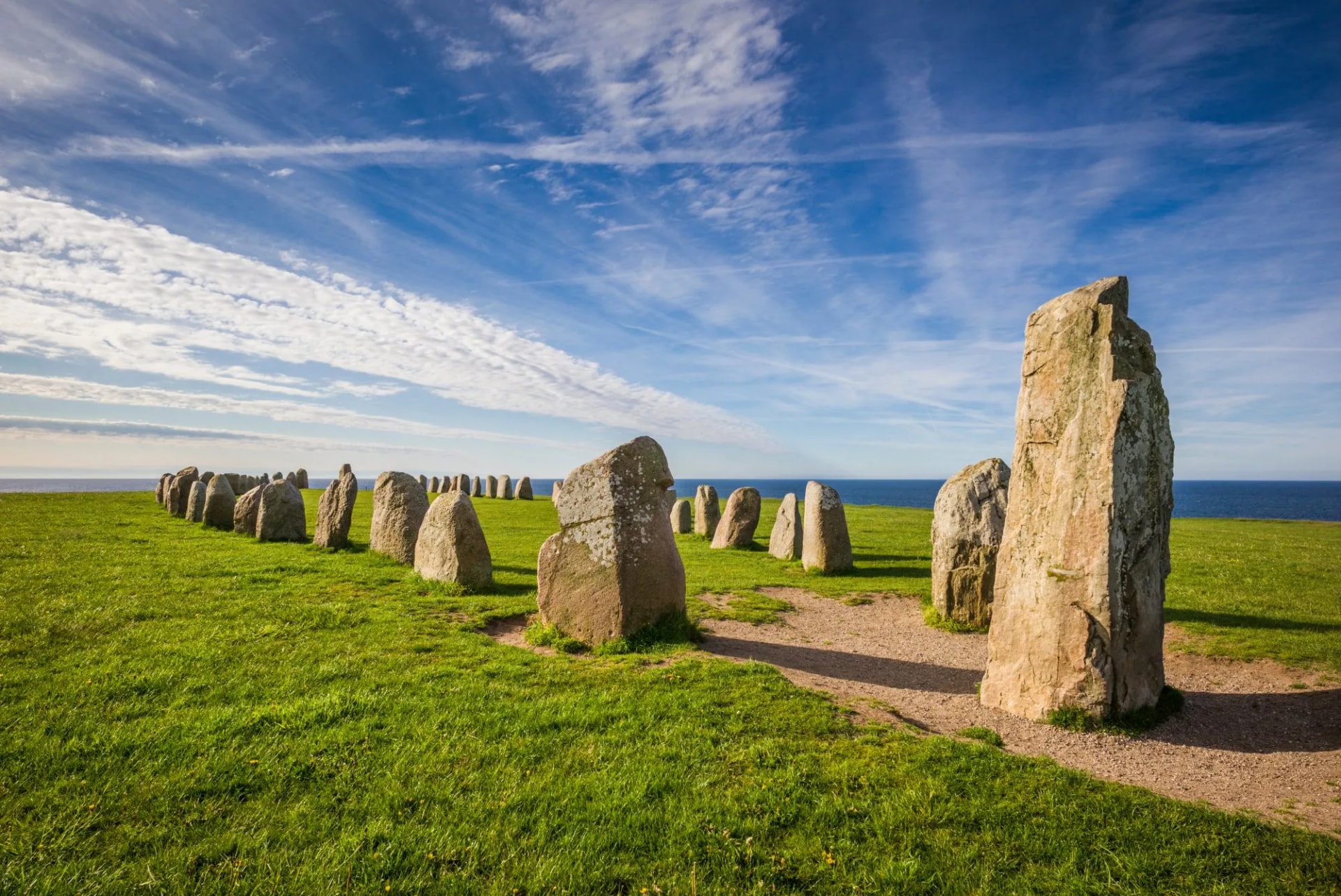 Ales Stenar stone ship setting on green grass overlooking the sea under a blue sky.