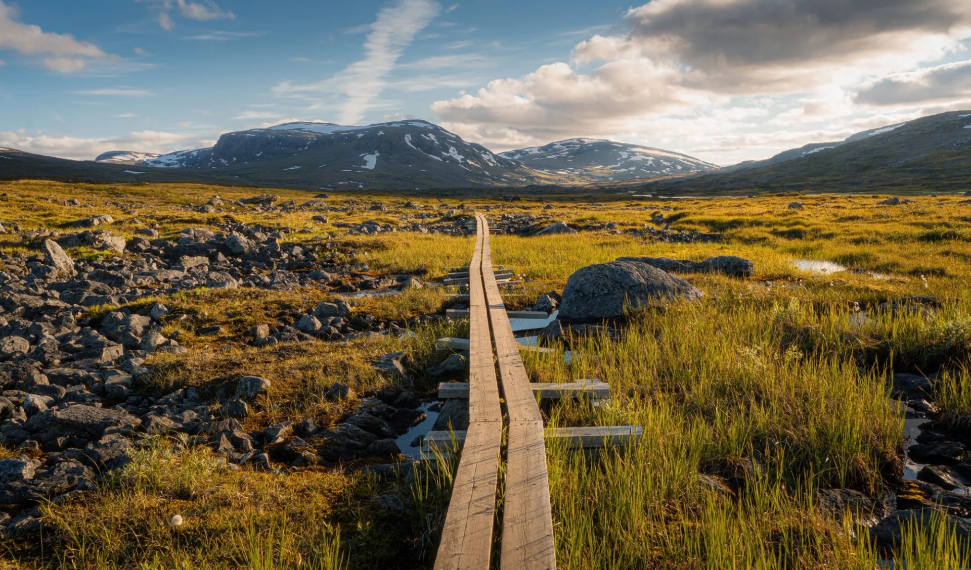Wooden path across marshland toward snow-capped mountains on Kungsleden trail in Lapland.
