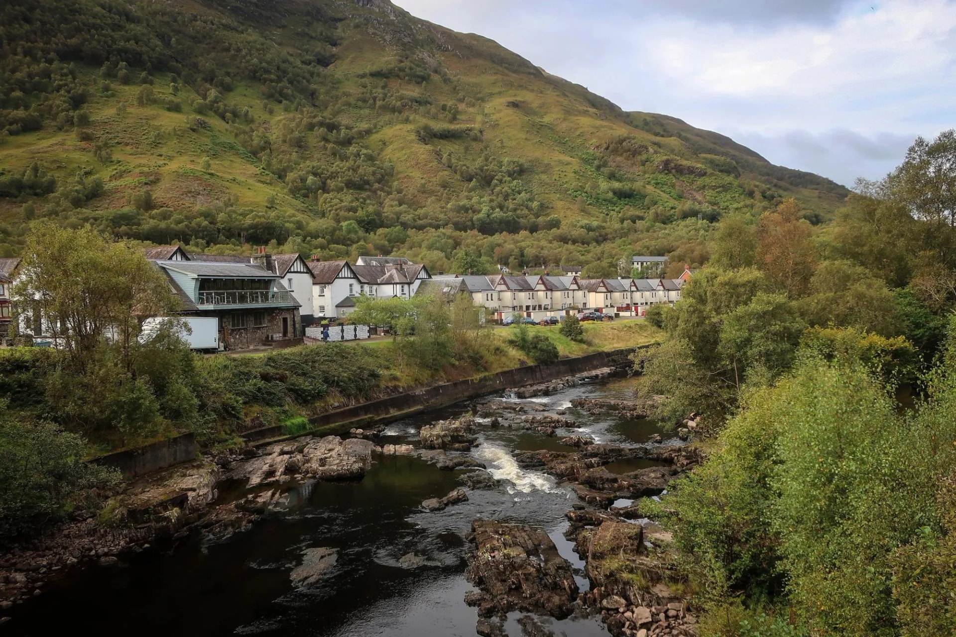 Rocky river rapids flowing past white houses nestled at the base of a large, green, forested mountain.
