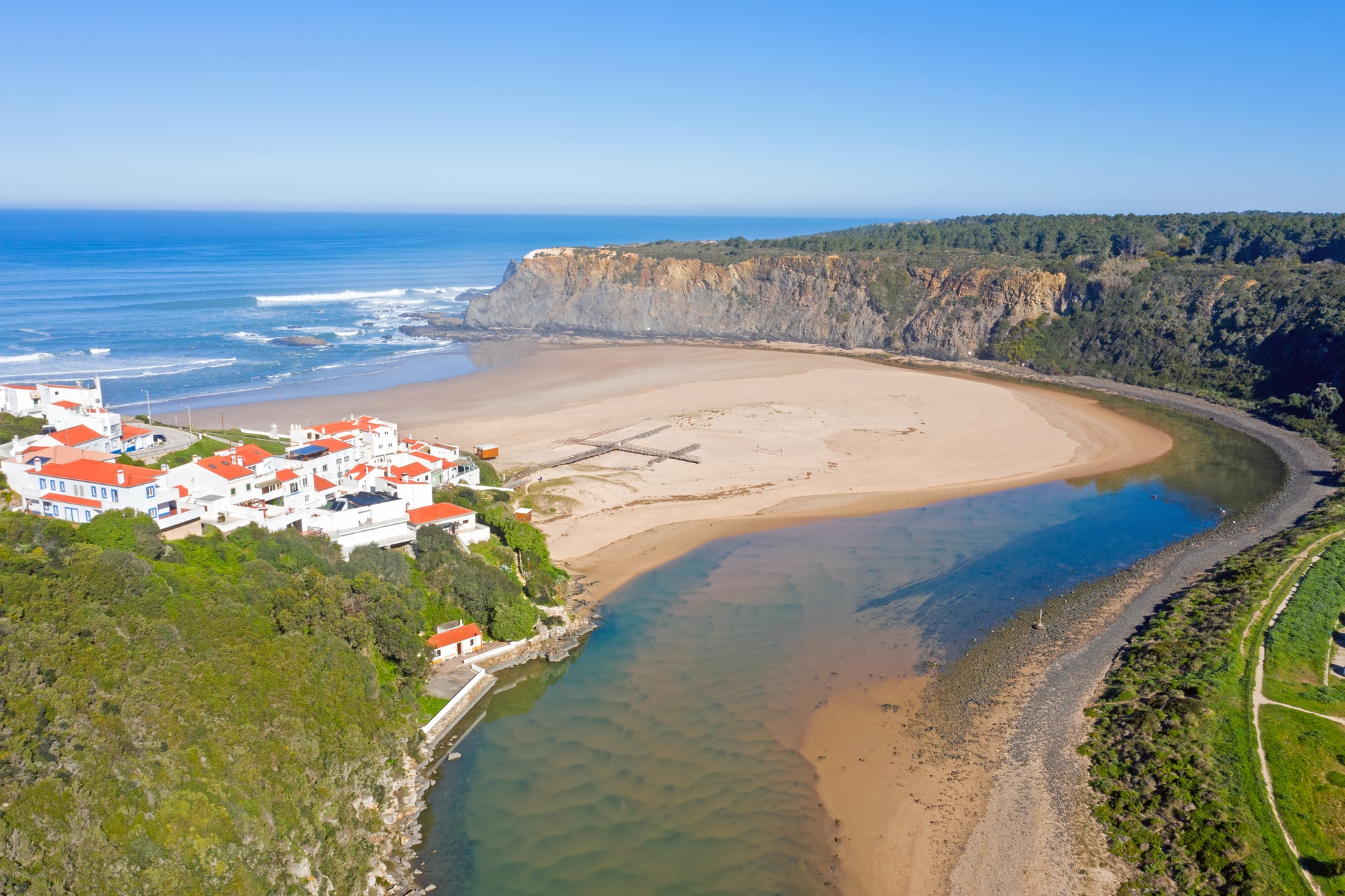 Aerial view of Praia de Odeceixe with white houses, river meeting the ocean, and sandy beach.