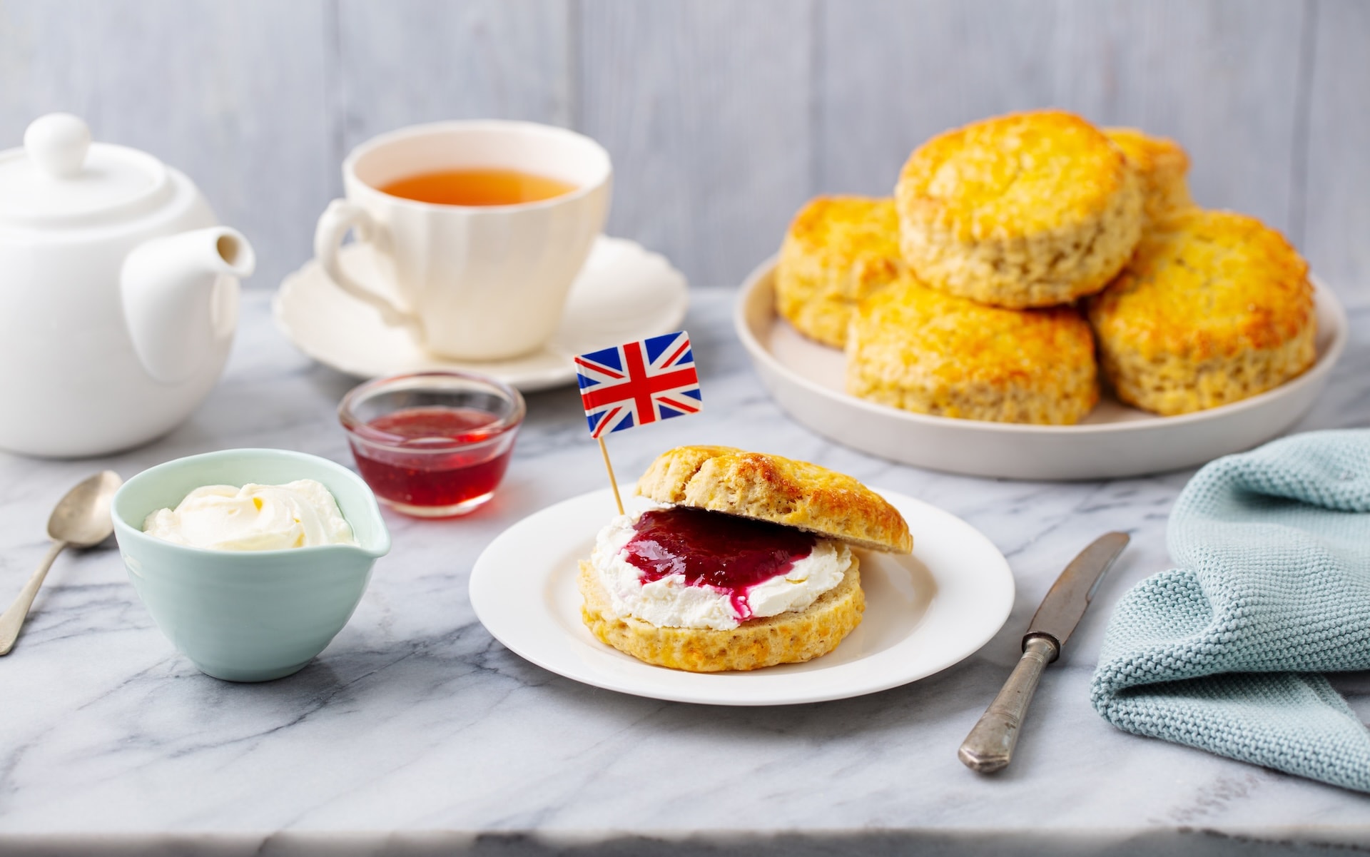 Scone with cream, jam, and Union Jack flag next to tea service on marble.