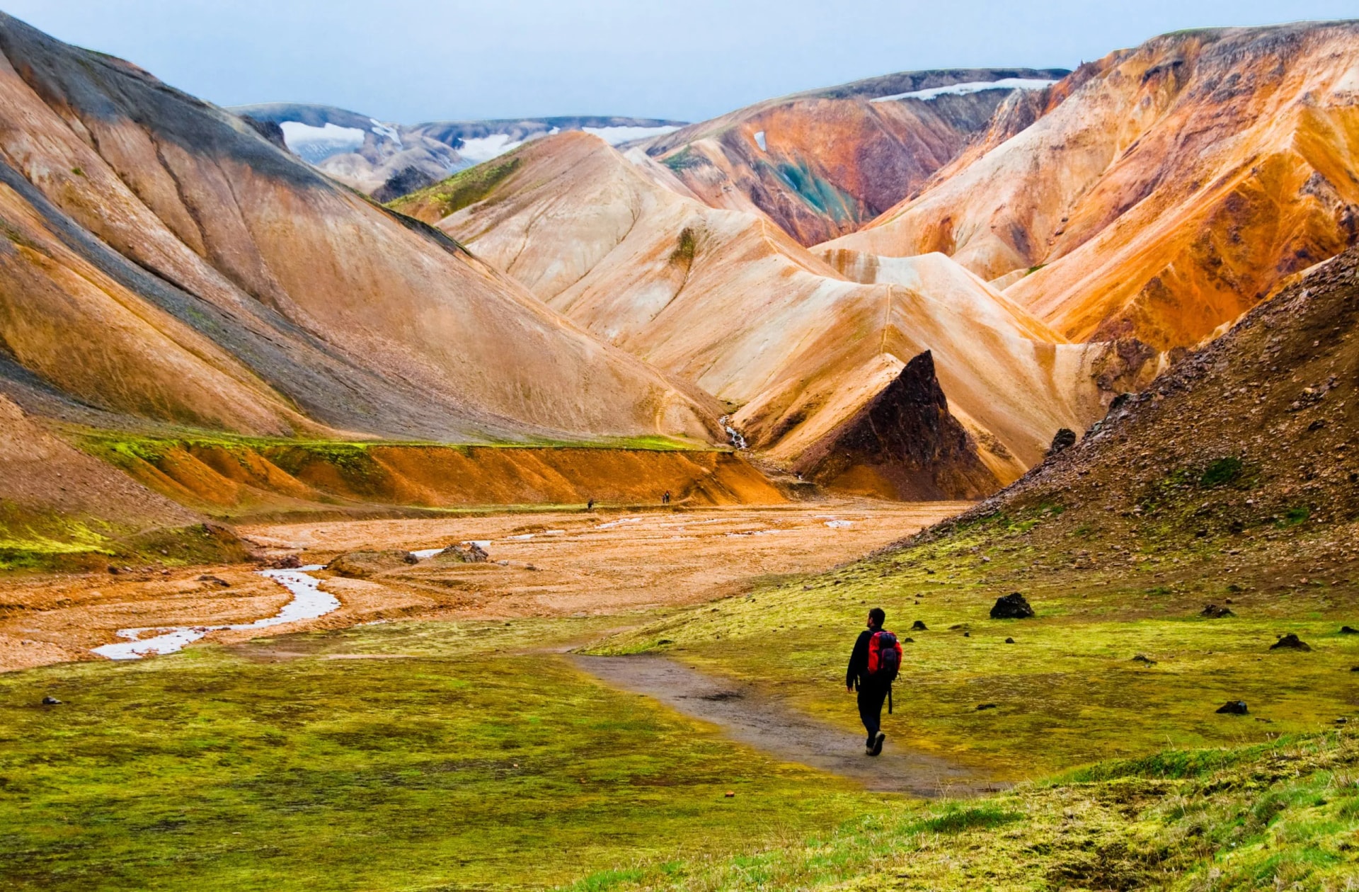 Hiker walking toward colorful rhyolite mountains and a stream in Landmannalaugar, Iceland.