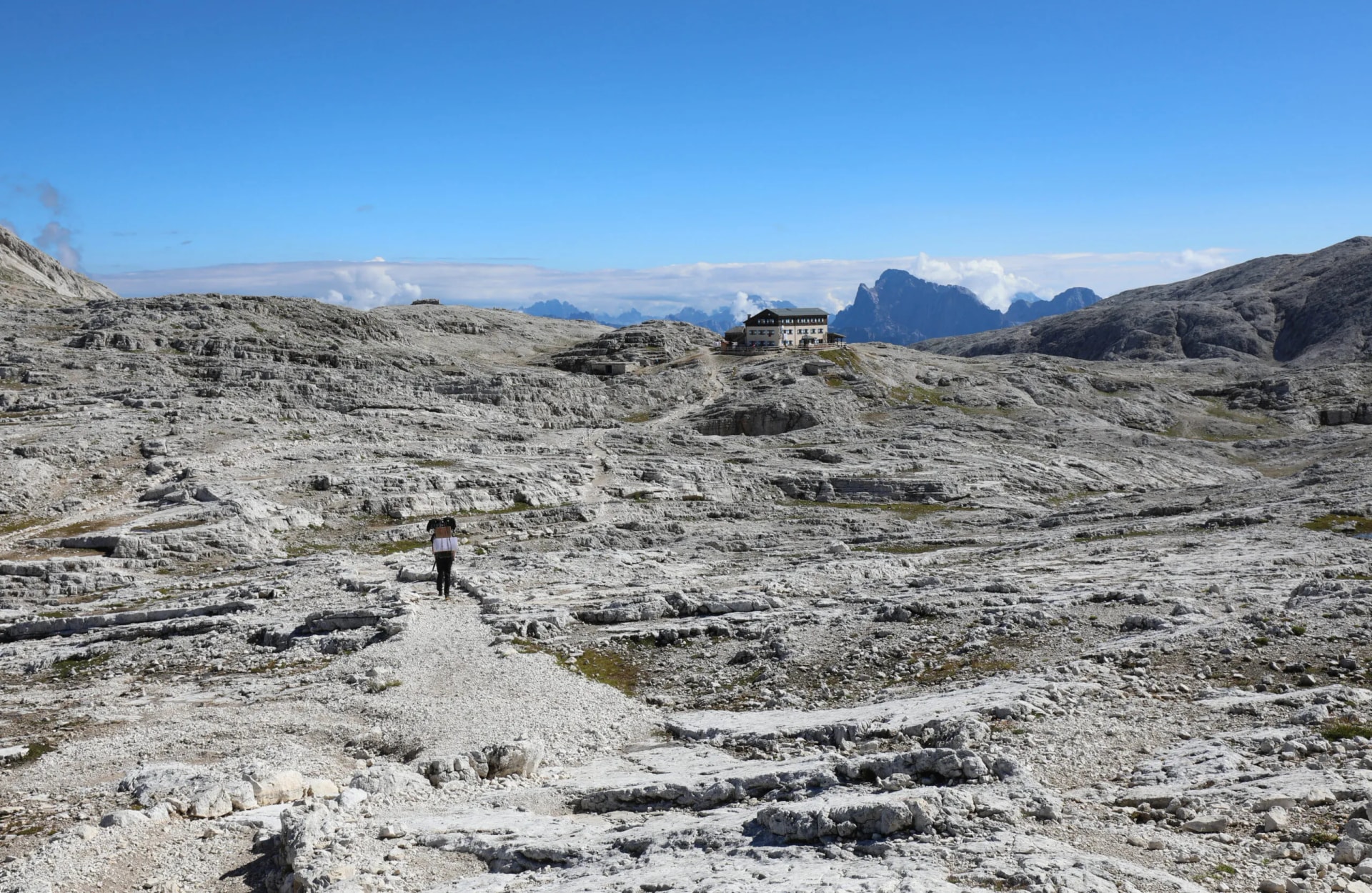 Hiker walking toward Rifugio Pale di San Martino across rocky alpine terrain.