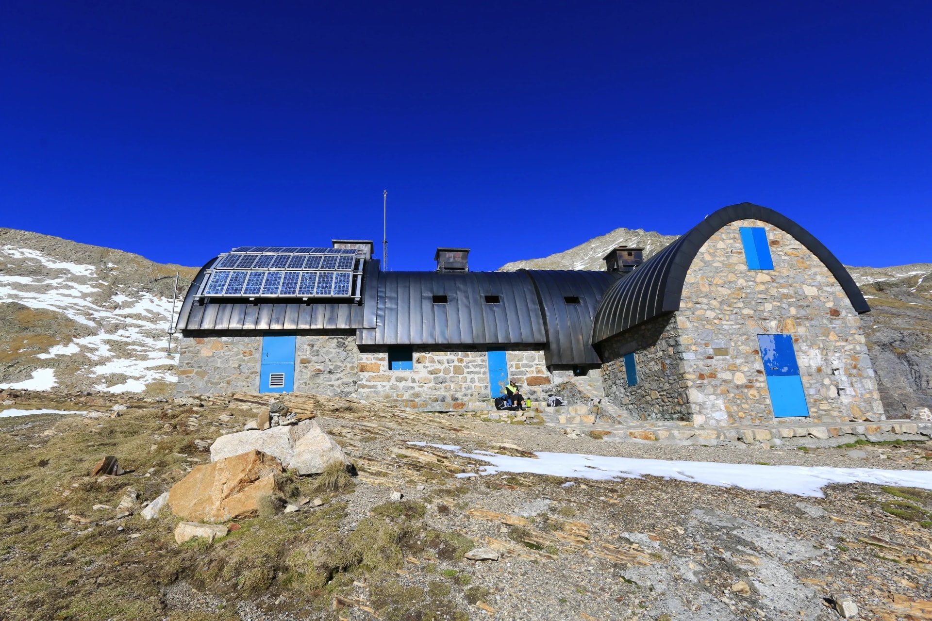 Stone mountain refuge with solar panels and blue doors under a clear blue sky.
