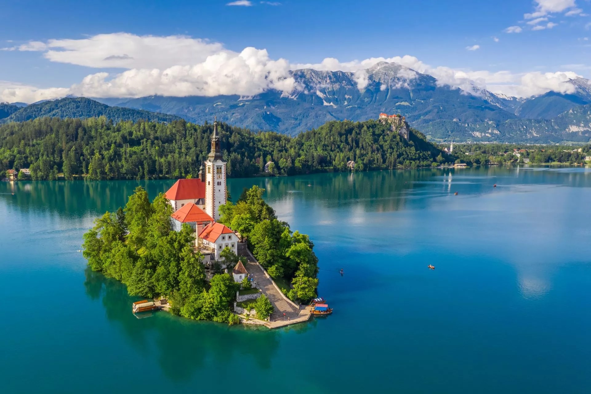 Lake Bled island church with boats on turquoise water and Julian Alps mountains.