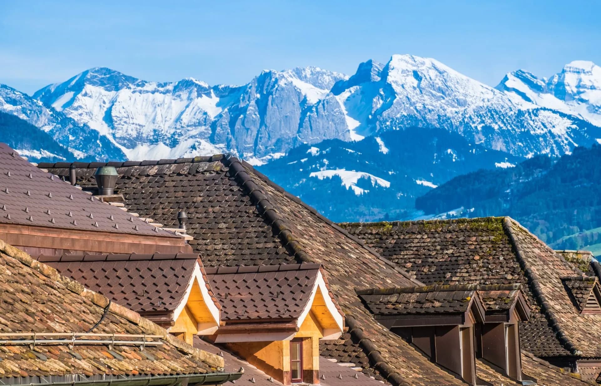 Rooftops in foreground with snowy Alpine mountains backdrop under clear blue sky