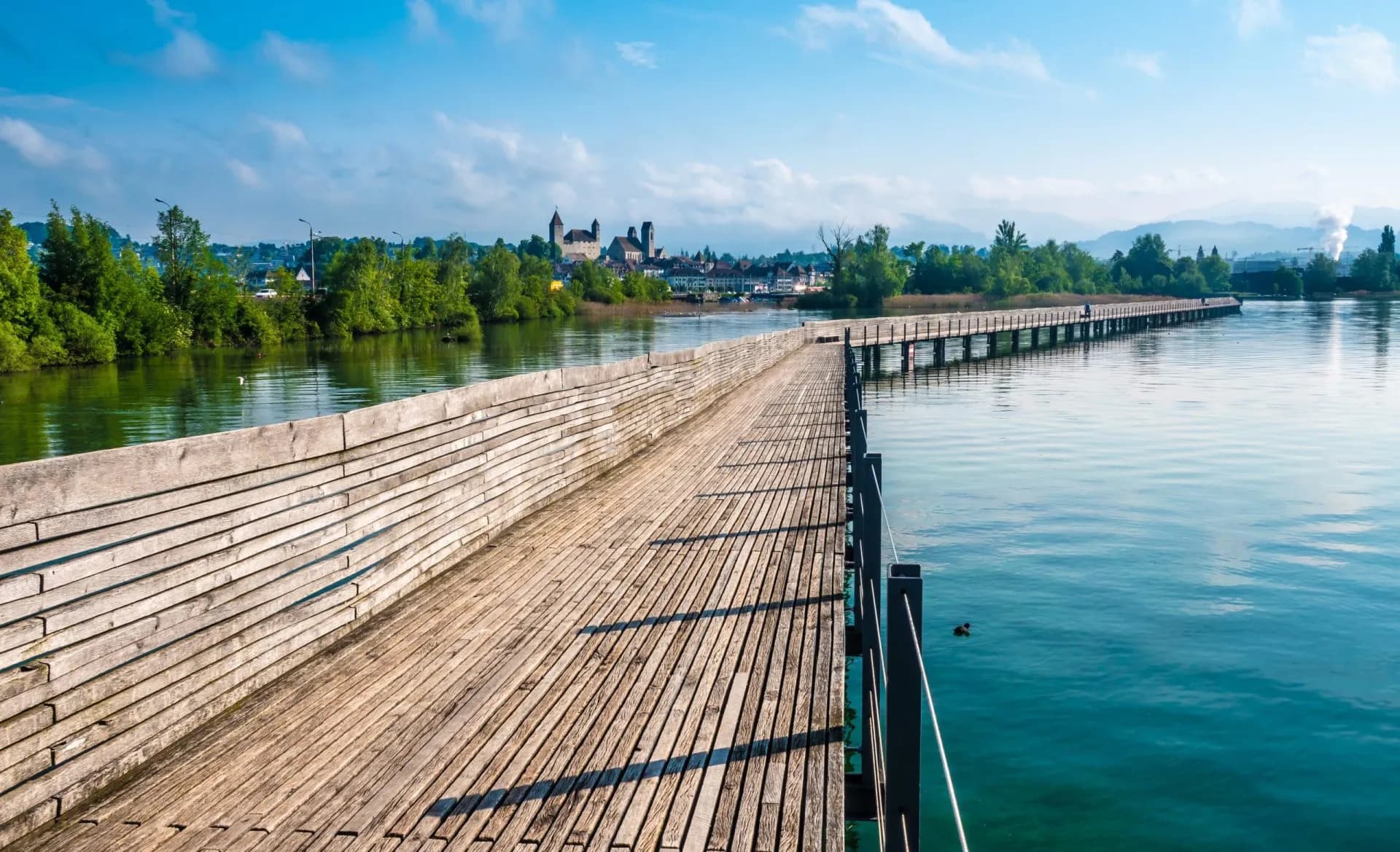 Wooden pedestrian bridge crossing Zurich Lake toward a town with a castle under a blue sky.