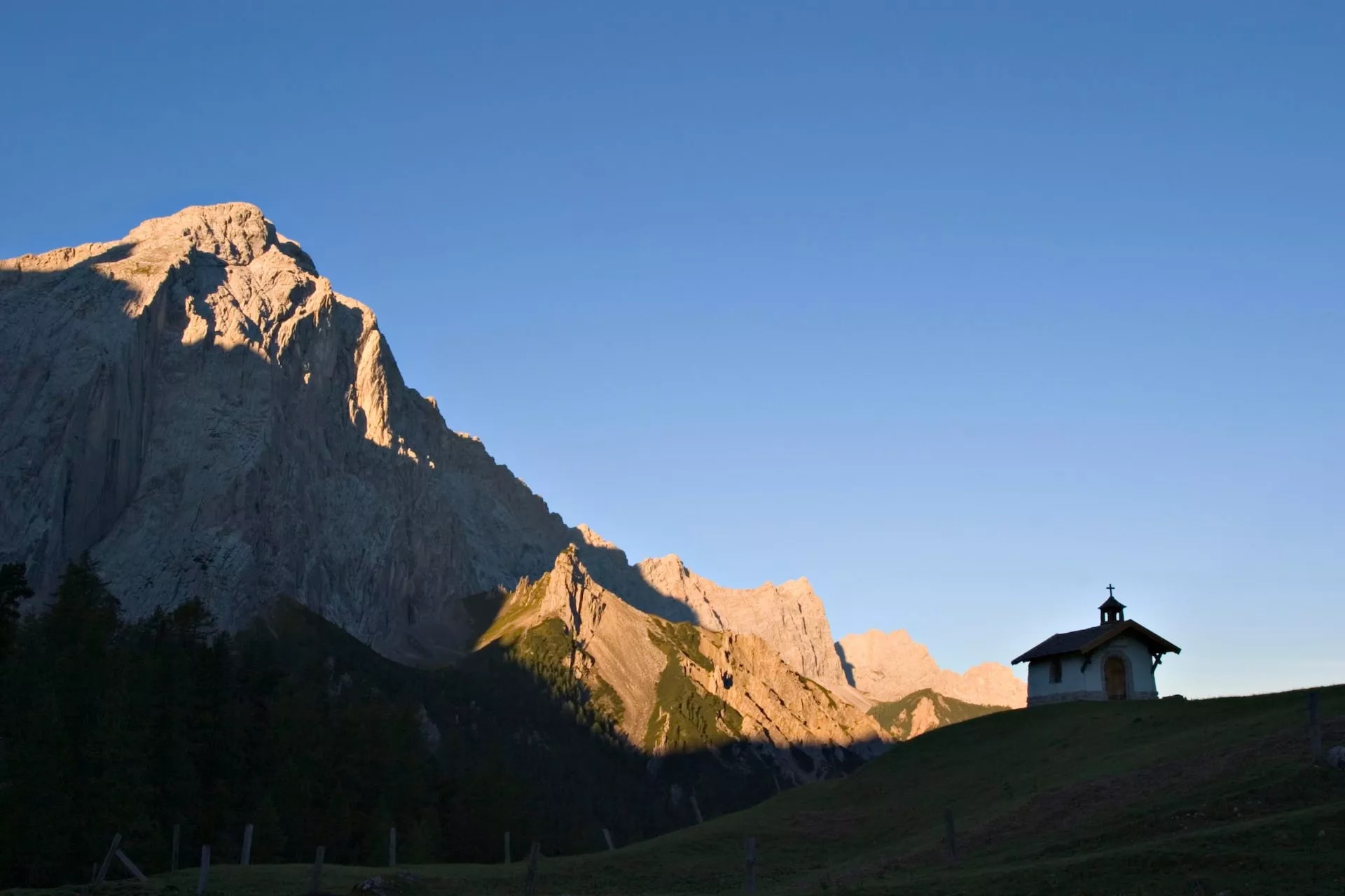 Small chapel on a grassy hill below sunlit alpine mountains under a clear blue sky.