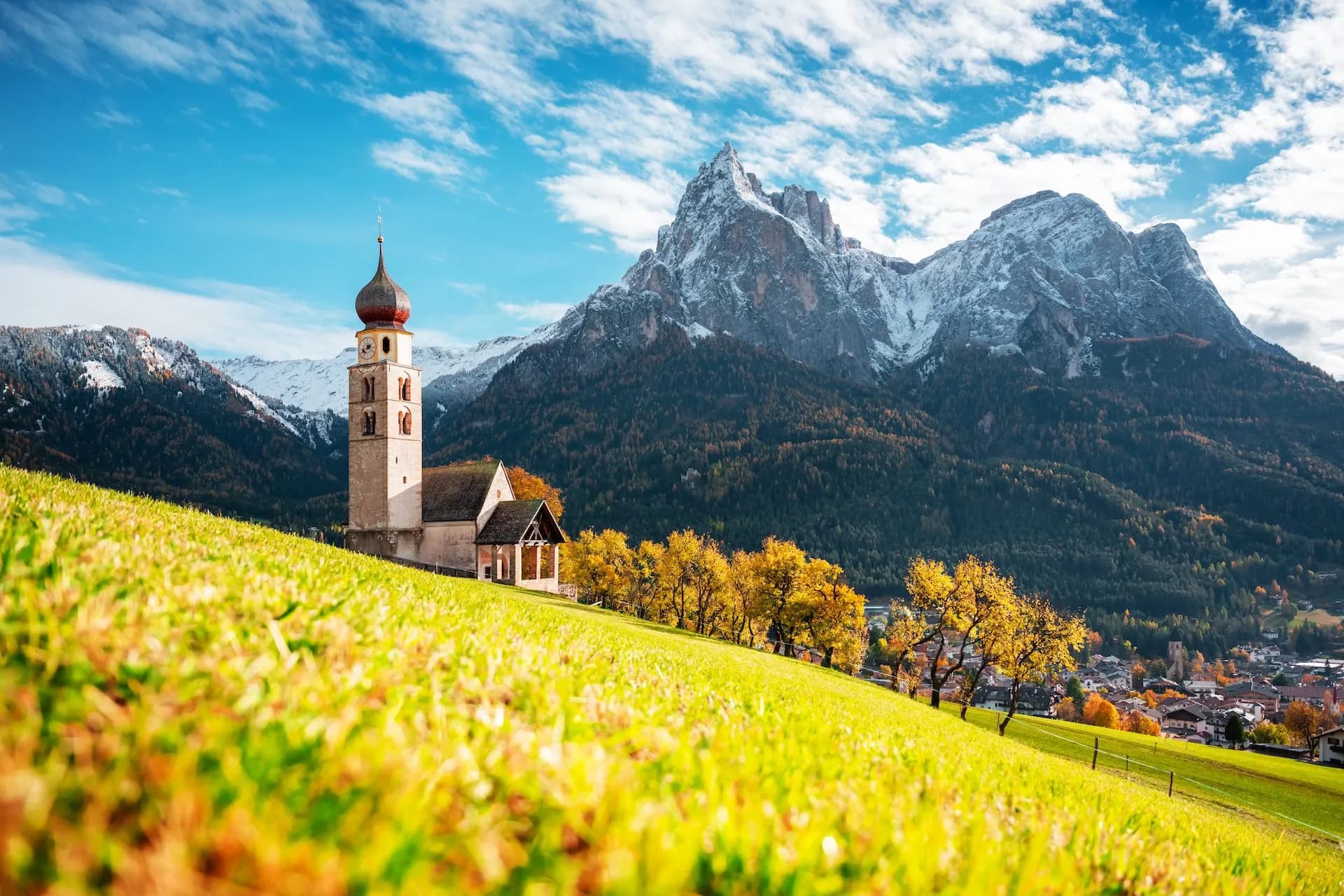 Church in Kastelruth on a grassy hill with snow-capped mountains and autumn trees.