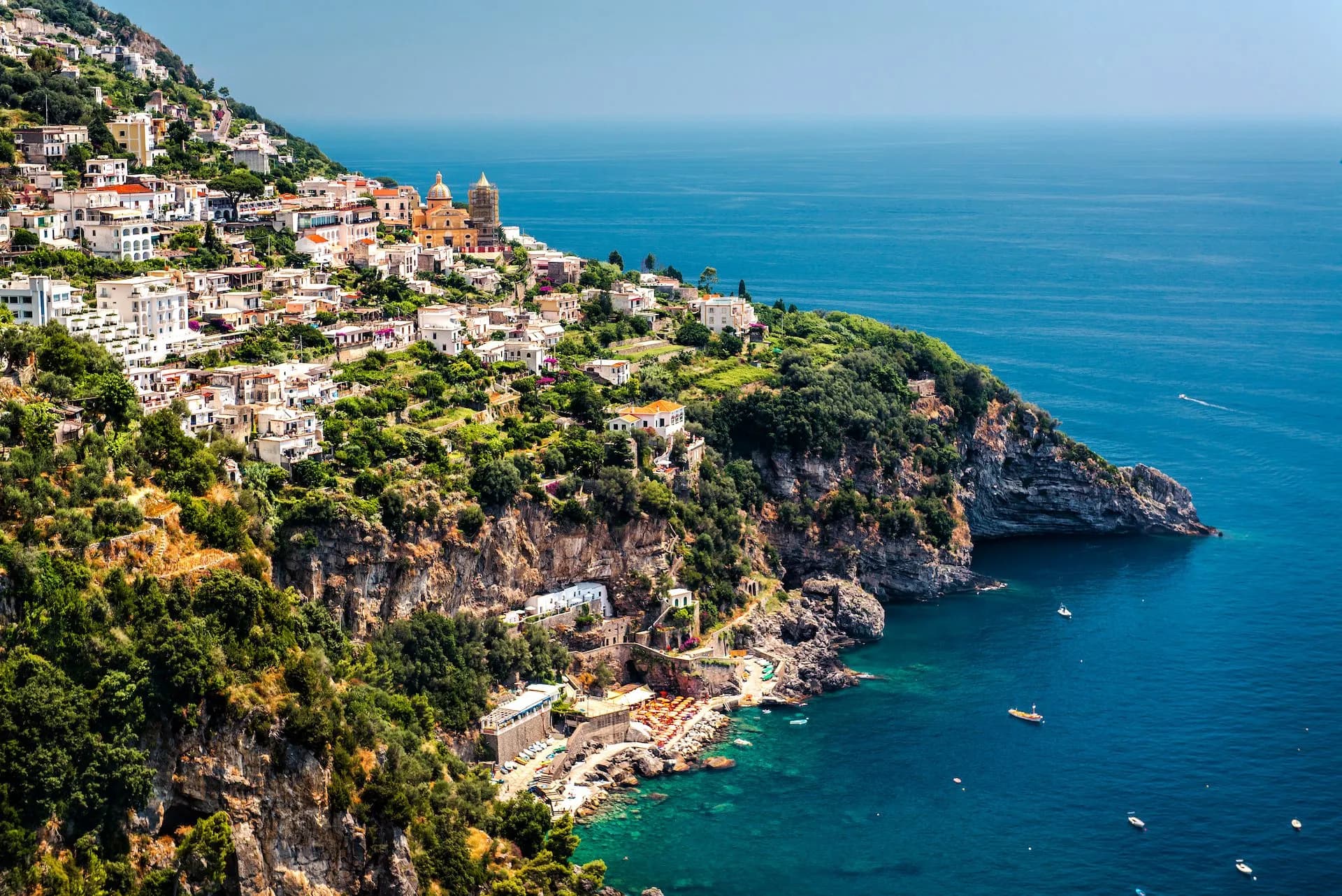 View of Praiano town built on steep green cliffs overlooking the deep blue sea with boats.